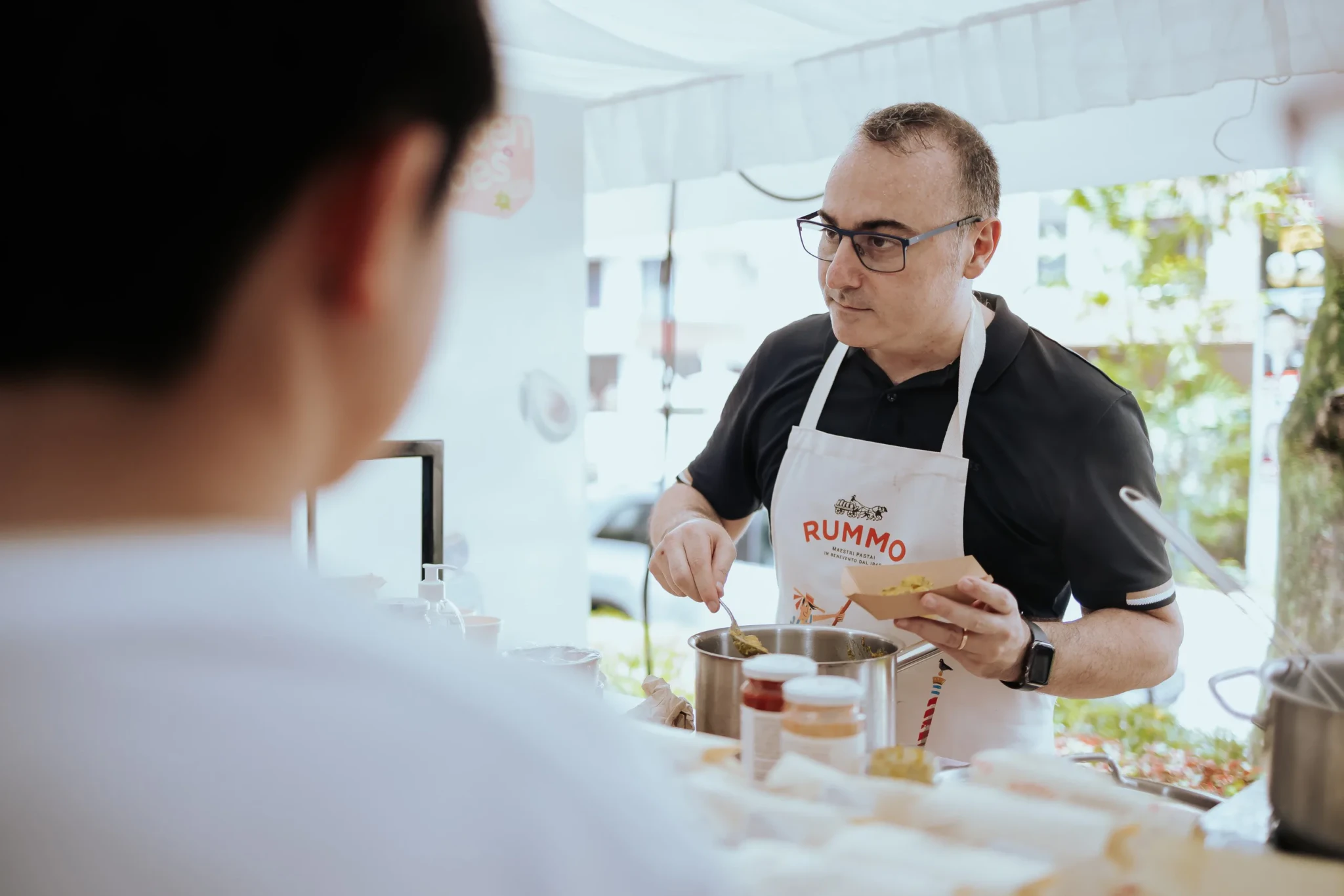 Man in black polo and RUMMO apron serving food from a pot at a pasta stall, with a customer in the foreground