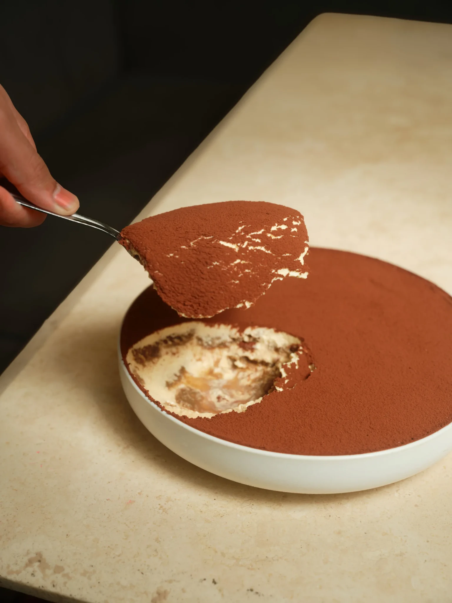 A spoon lifts a serving of cocoa-dusted tiramisu from a white bowl on a stone-look table