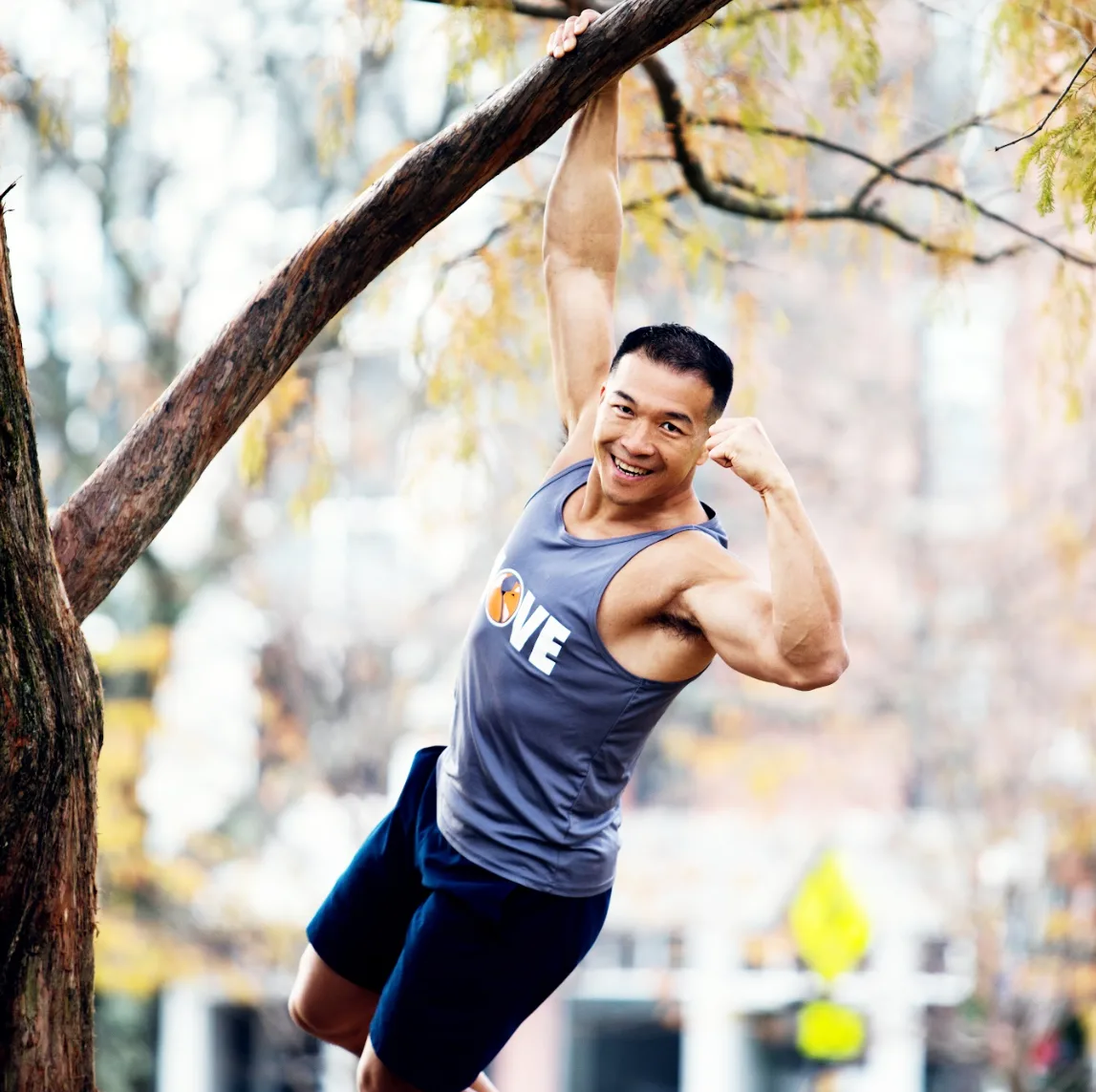 Jimmy Choi hanging from a tree branch in a park while flexing his arm
