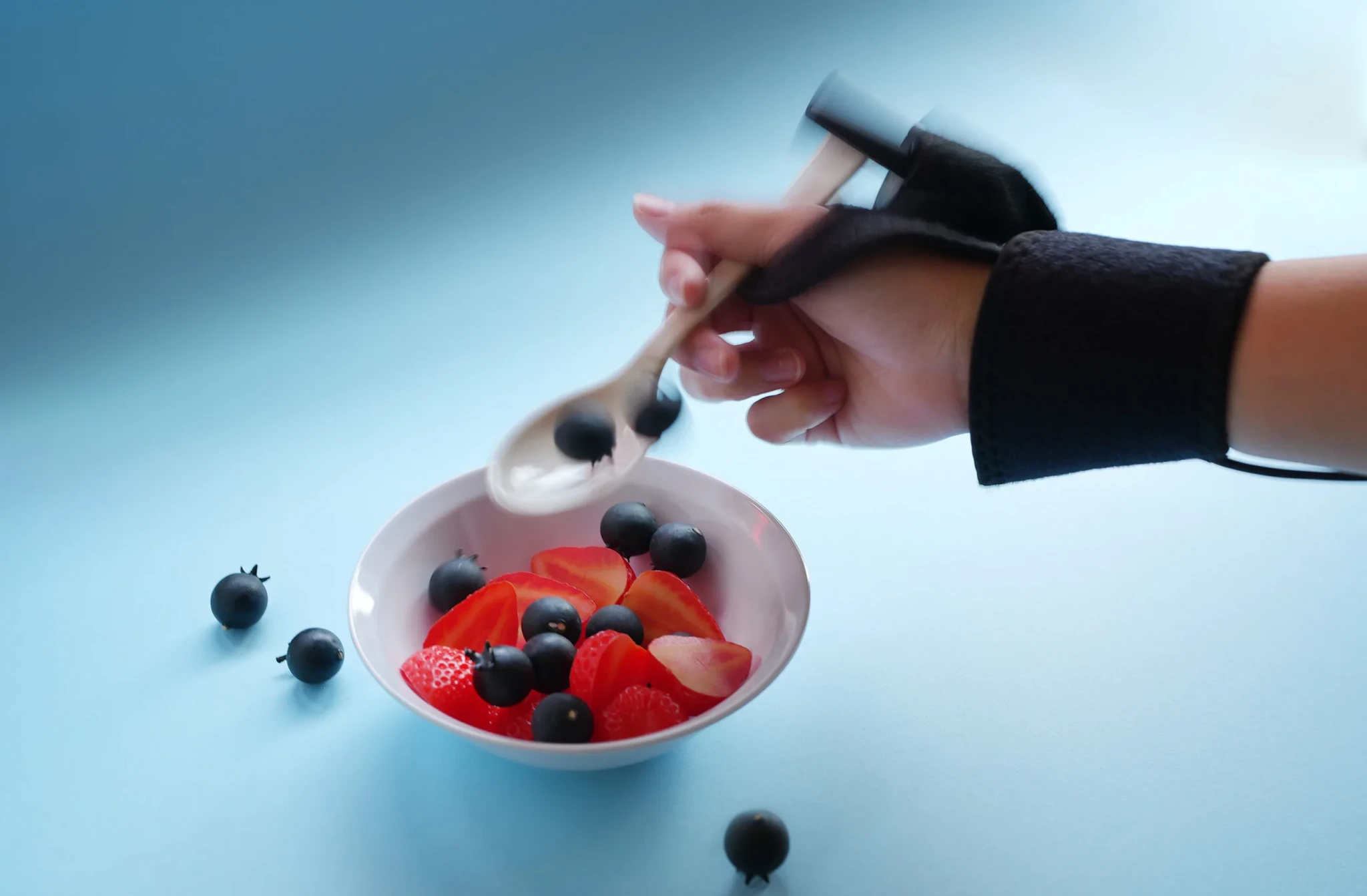 A hand in a black support splint lifting blueberries with a spoon from a bowl of berries