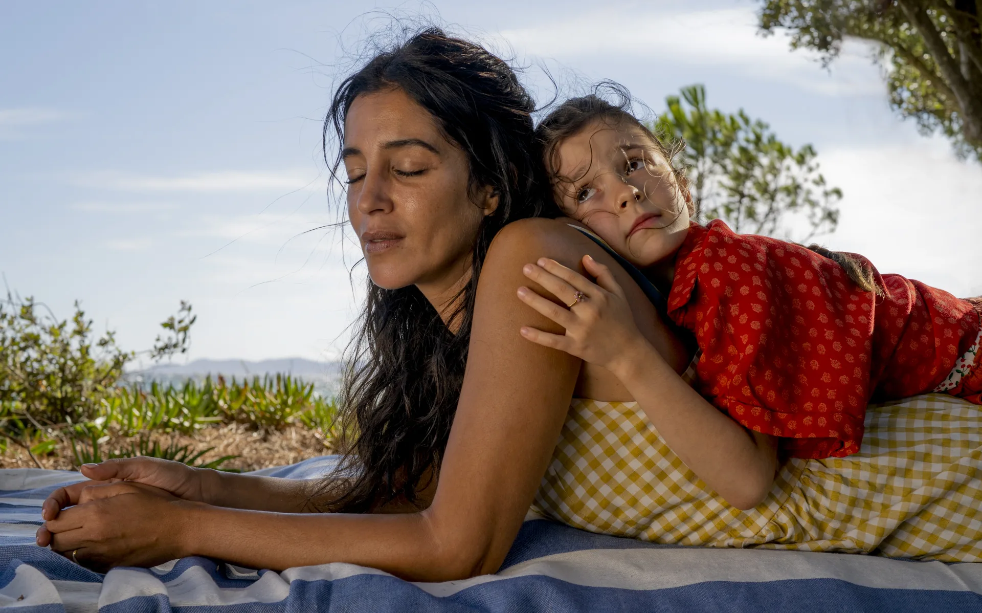 A woman with long dark hair lies on a blue-and-white striped blanket as a young girl rests her head on her shoulder