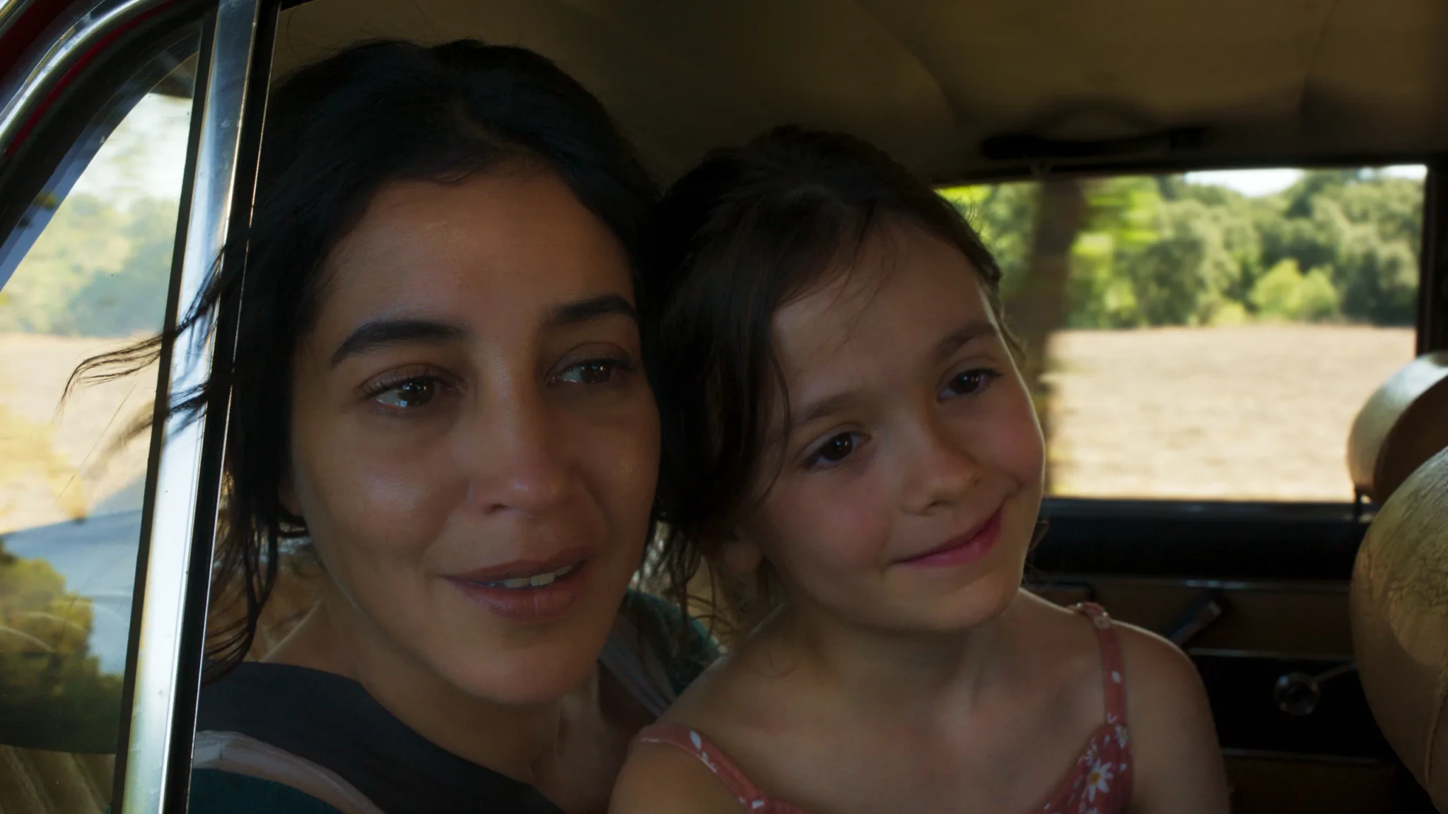 Woman and young girl sitting together in the back seat of a car, looking out the window at sunlit fields and trees