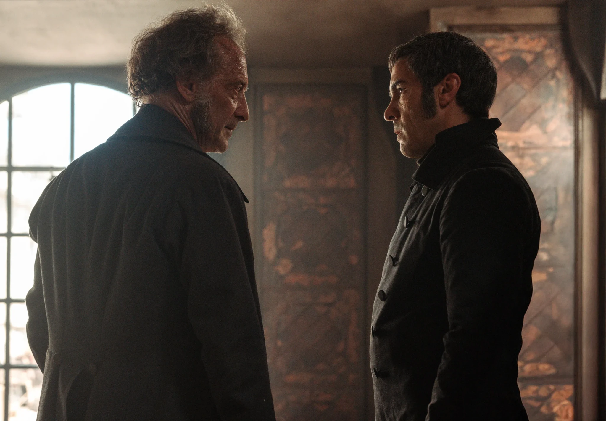 Two men in dark period coats face each other in a dim interior beside a barred arched window