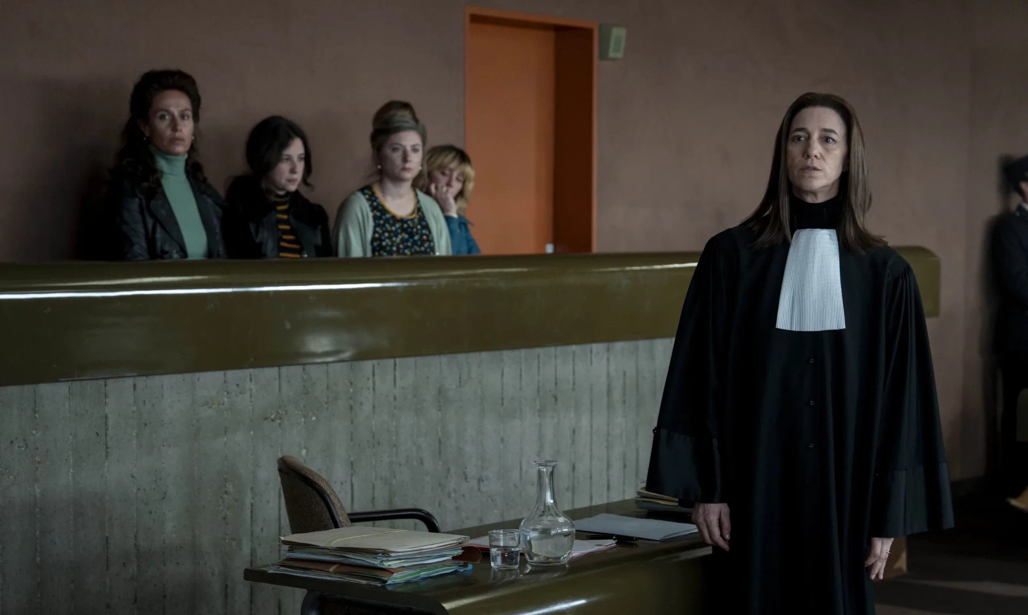 A lawyer in a black robe and white bands stands in a courtroom as women sit behind the defence bench