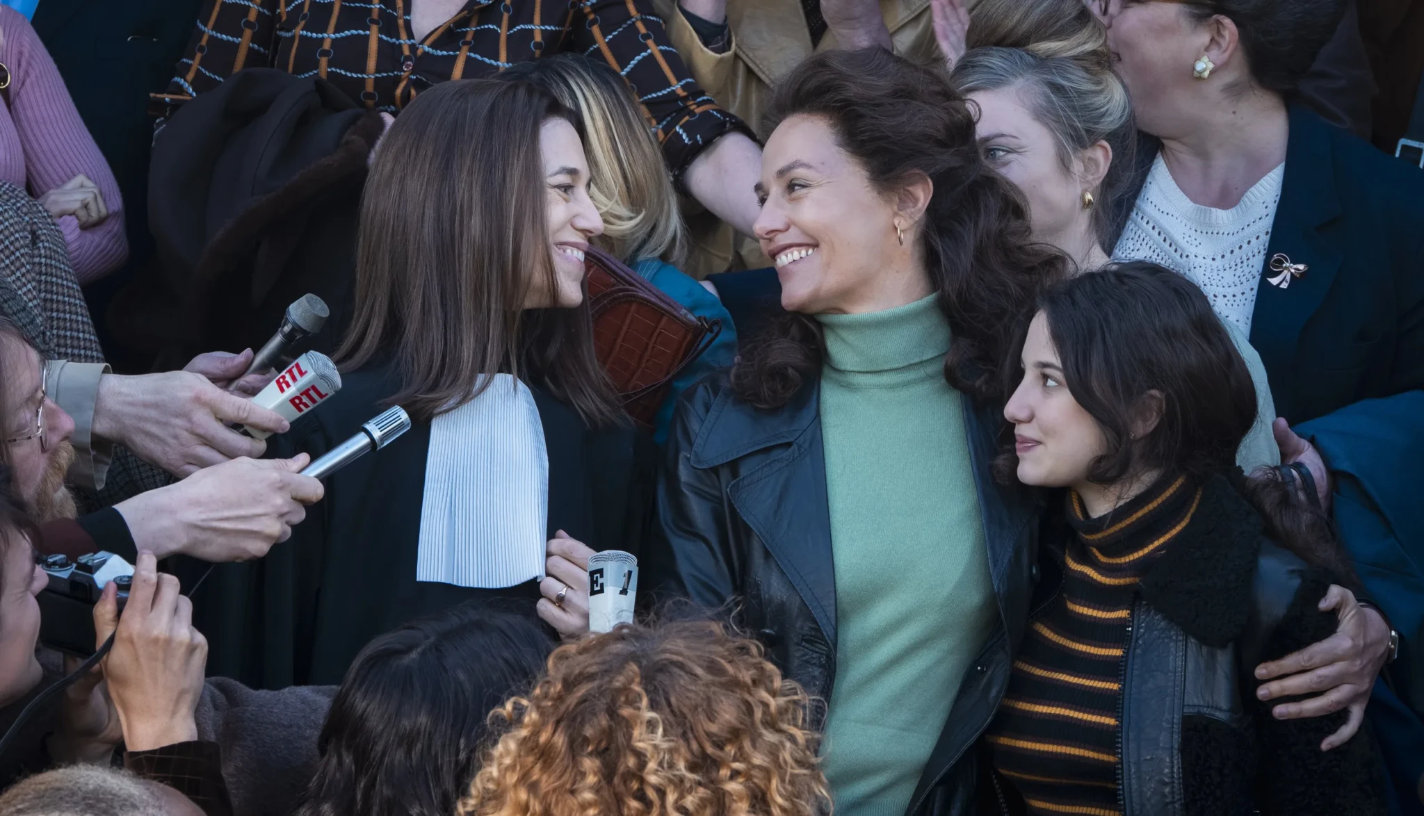 Smiling women in a crowd with reporters' microphones, one wearing black lawyer's robes with a white jabot.