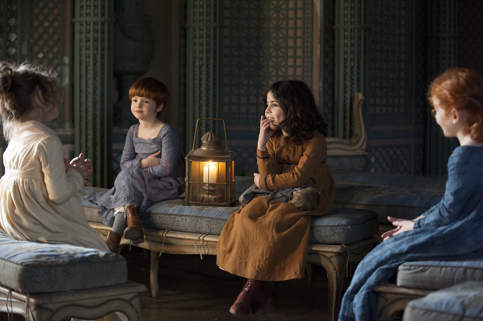 Four young girls in period costume sit around a lantern on an ornate blue sofa in a dim room with green lattice screens.