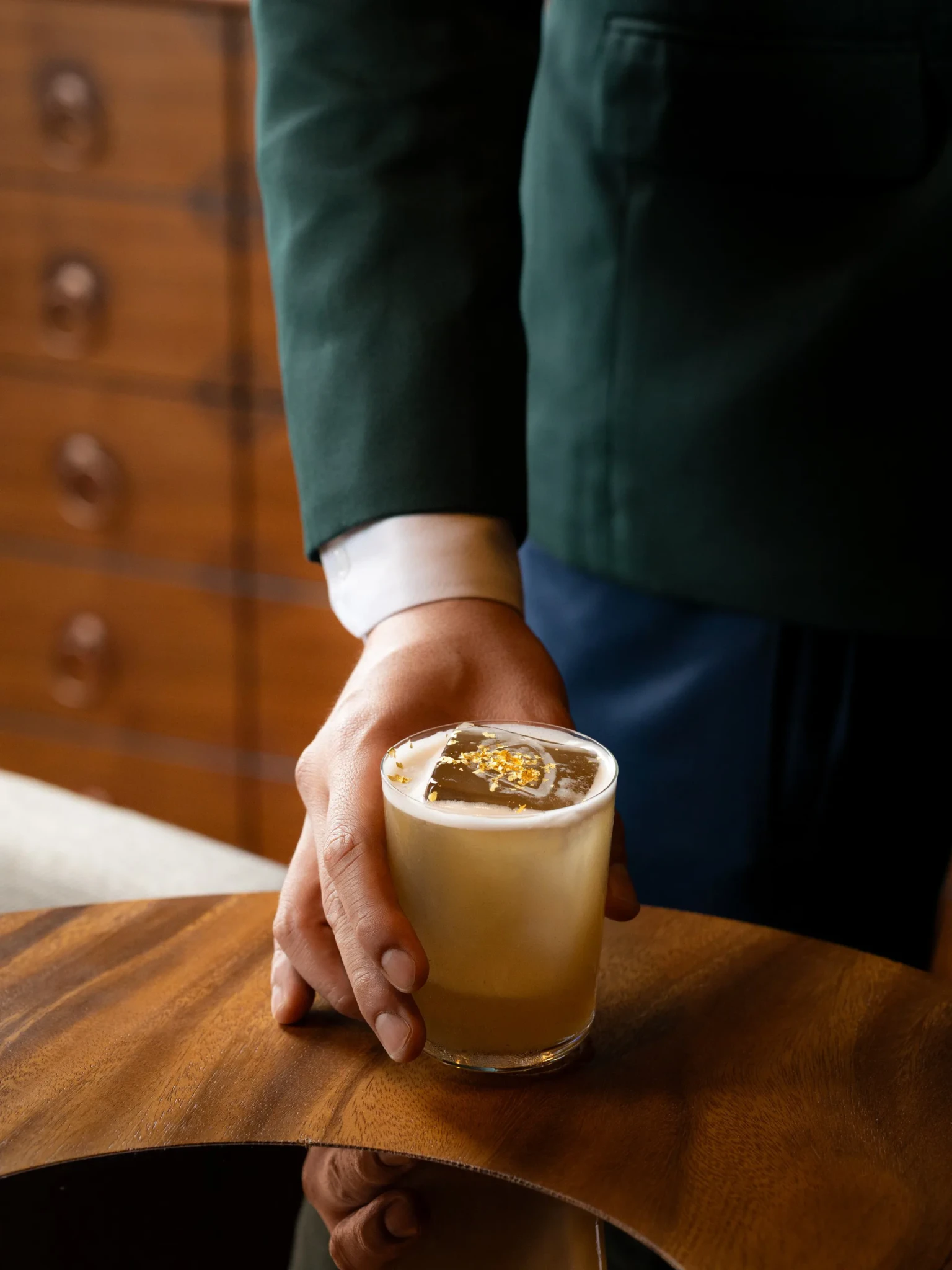 A frothy cocktail with a large clear ice block and gold leaf flakes held over a wooden bar