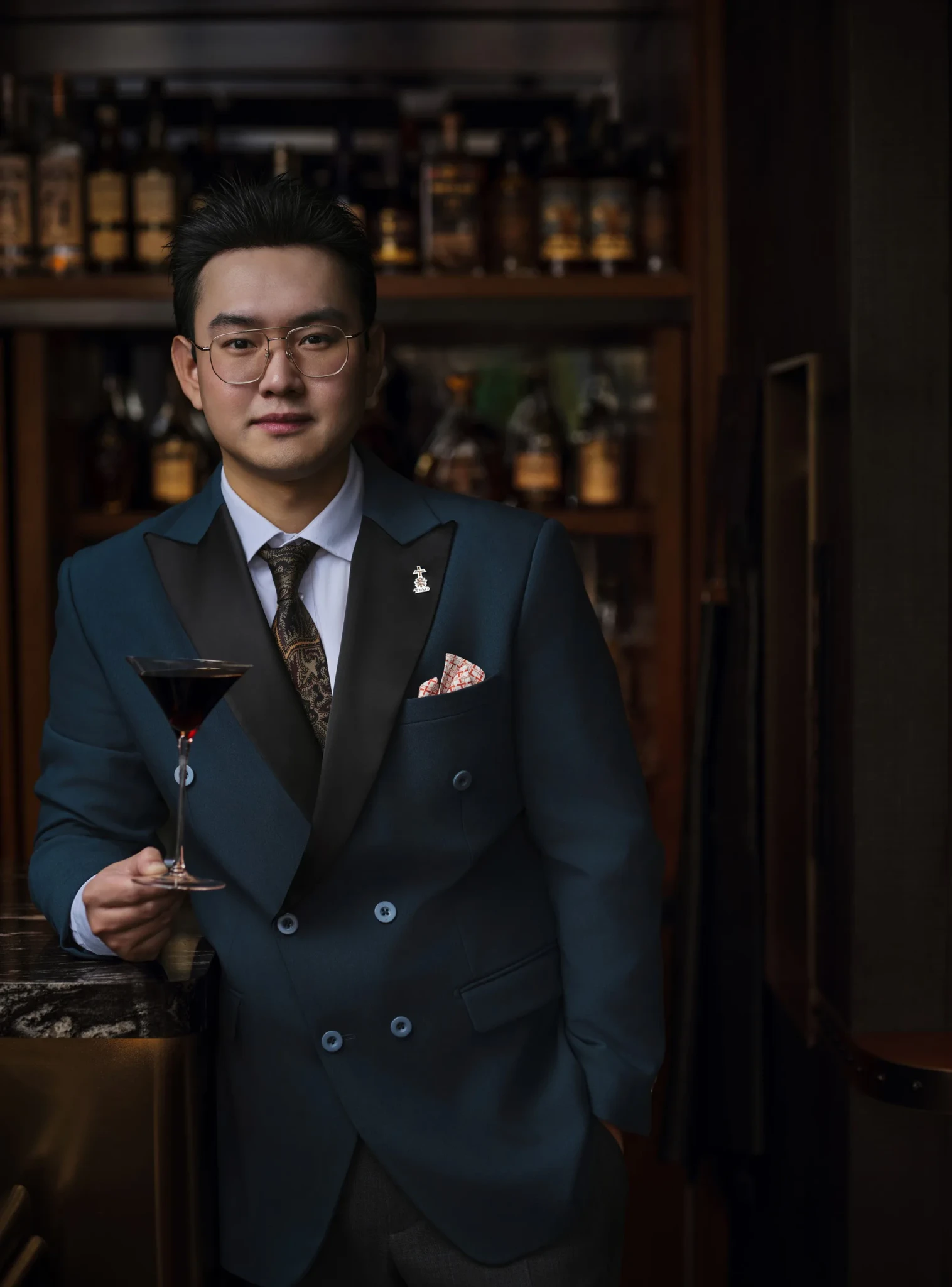Man in teal double-breasted suit and glasses holding a dark cocktail in a stemmed glass in front of a backbar of spirits.