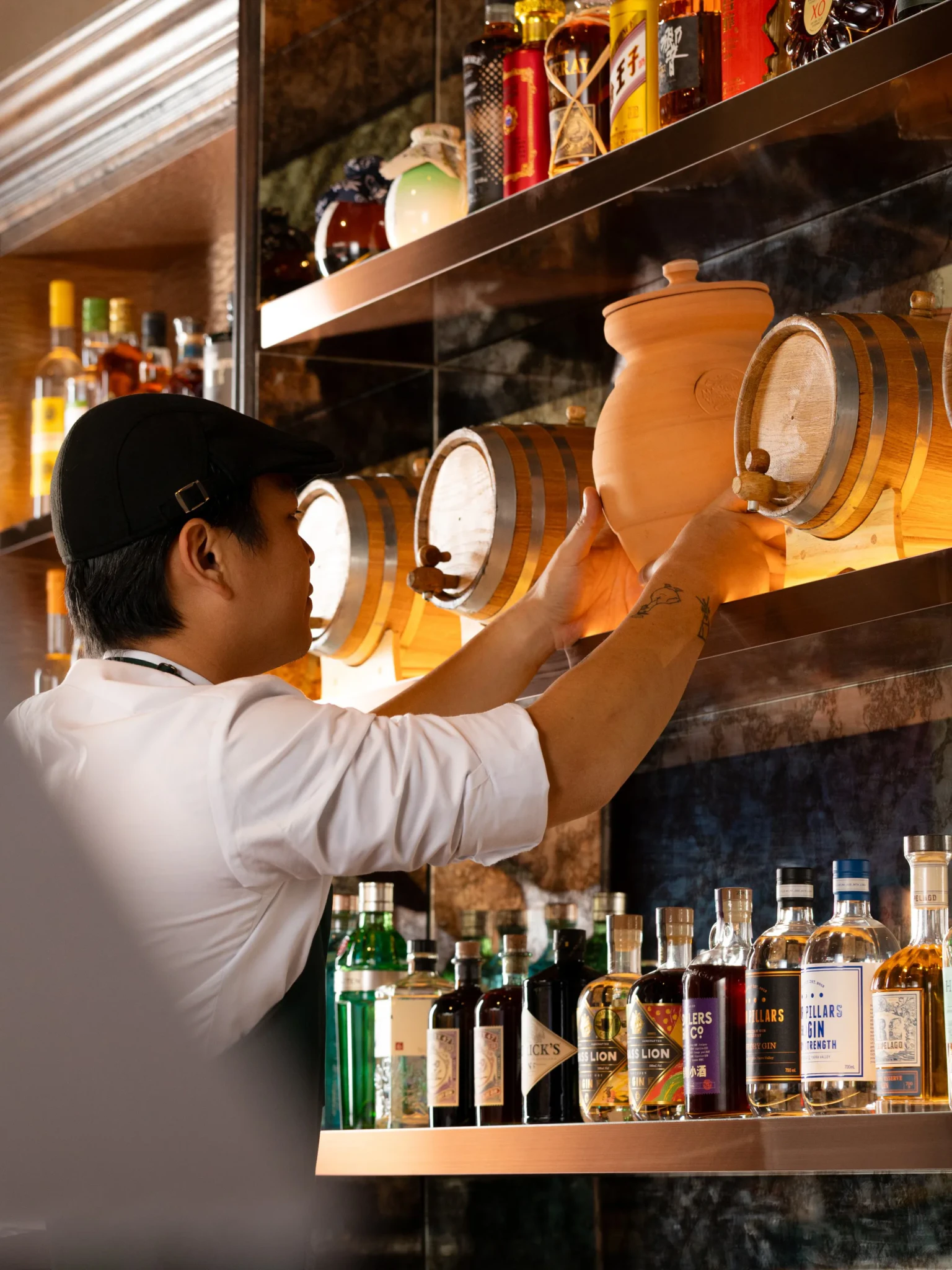 Bartender in a black cap placing a clay vessel on a shelf beside oak barrels and gin bottles at a bar