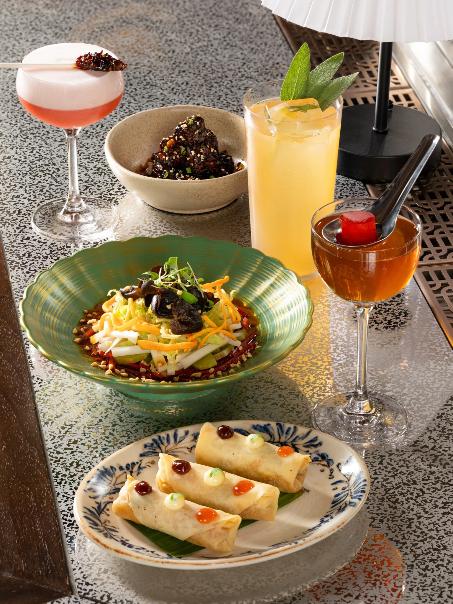 Spread of cocktails and small plates on a speckled bar table: pink coupe, highball with sage, amber drink, salad bowl and spring rolls.