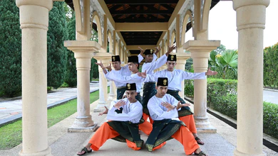 Six male dancers in white baju, green sampin and orange trousers and songkok strike poses between stone columns in an arched walkway.