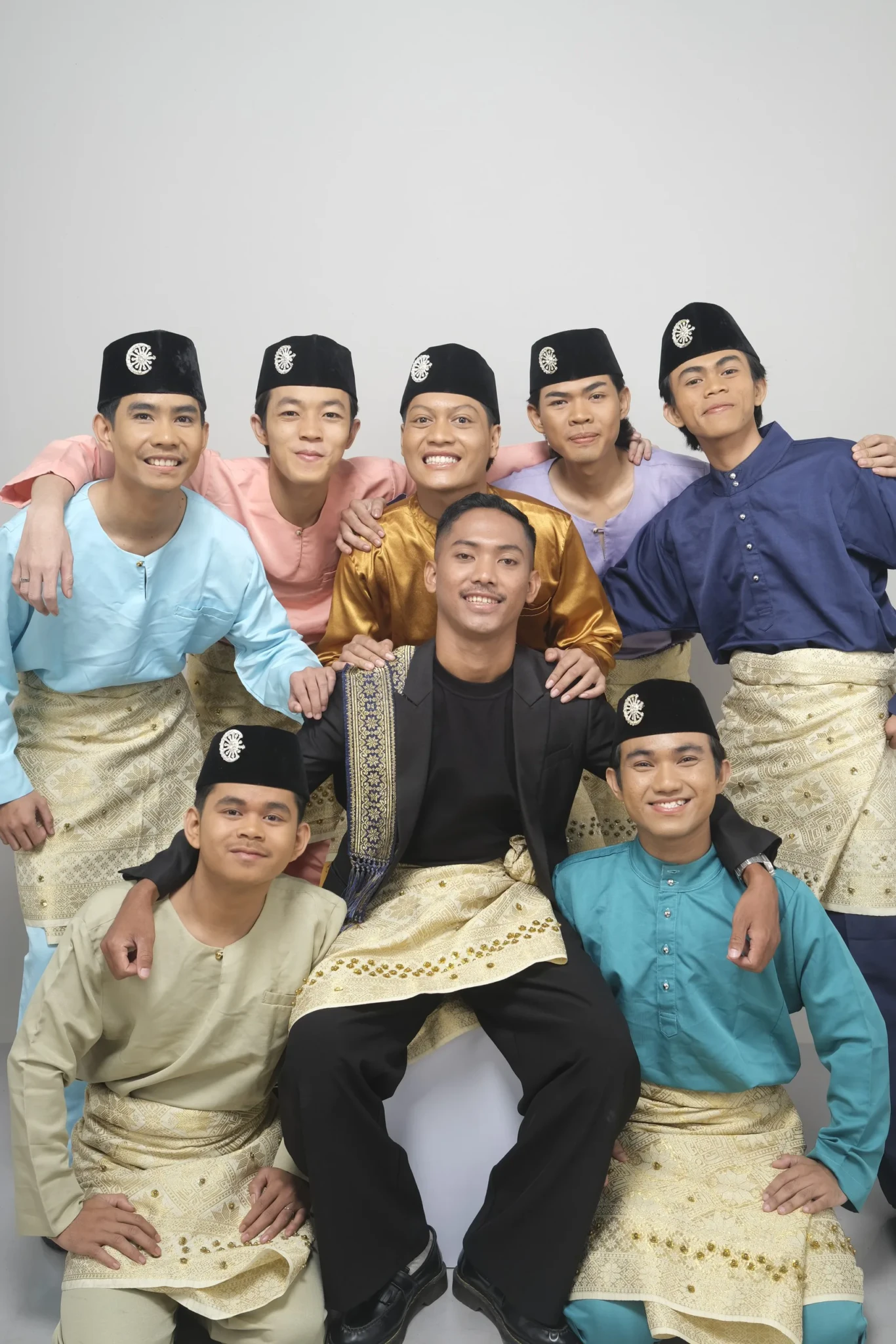 Nine smiling men in colourful traditional Malay baju Melayu and black songkok posing together in a studio
