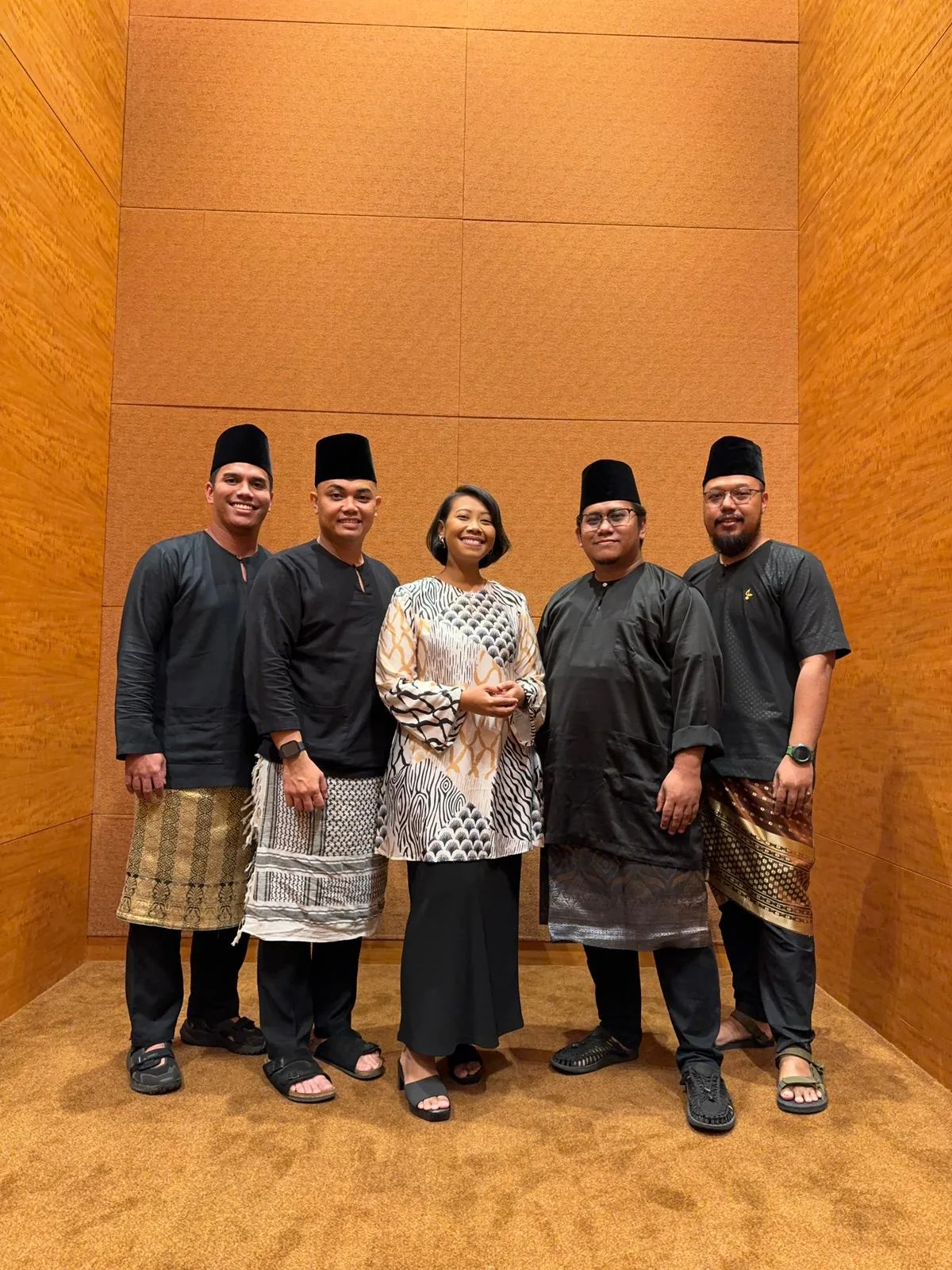 Five people in black songkok caps and baju Melayu with songket sampin posing together in a wood-panelled room