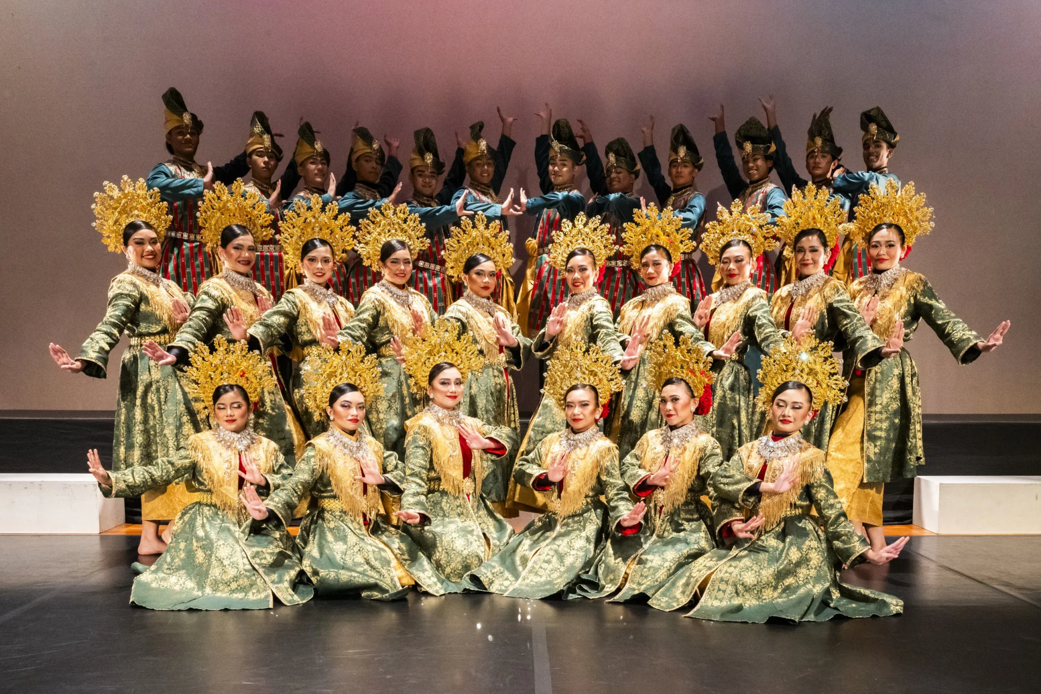 Group of performers in green and gold traditional Malay costumes with golden headdresses posing on stage in formation