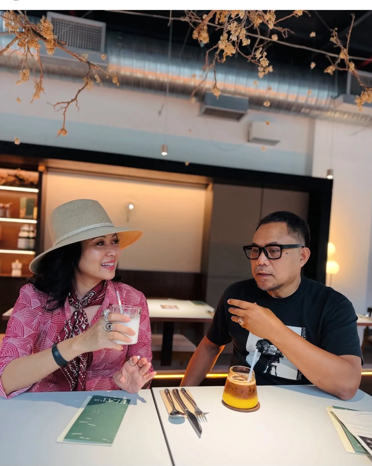 Wahyu Rahman in a black graphic T-shirt and glasses gesturing as he talks at a restaurant table with a companion in a wide-brimmed hat