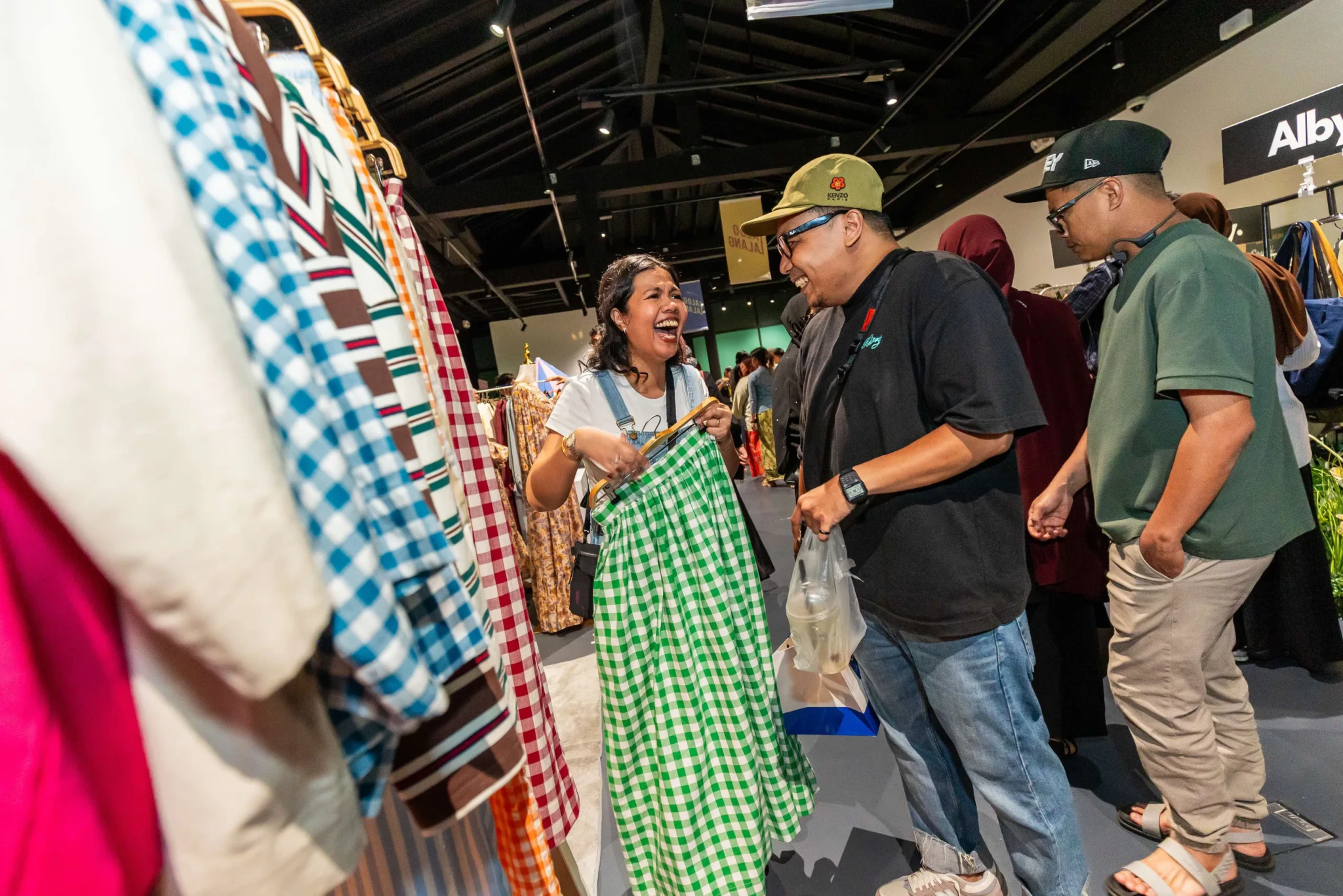 Visitors browsing gingham and striped garments on clothing racks, with a woman laughing as she shows a green gingham skirt to a companion