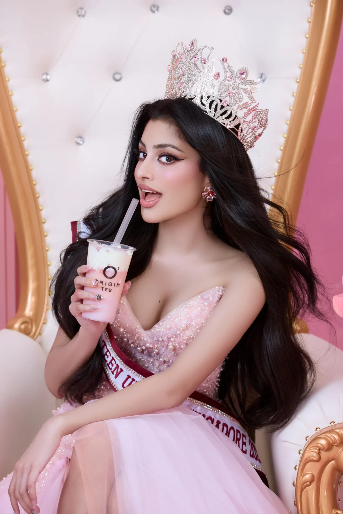 Woman in a pink embellished gown and tiara holding an Origin Tea drink with a straw, seated on a white and gold throne.