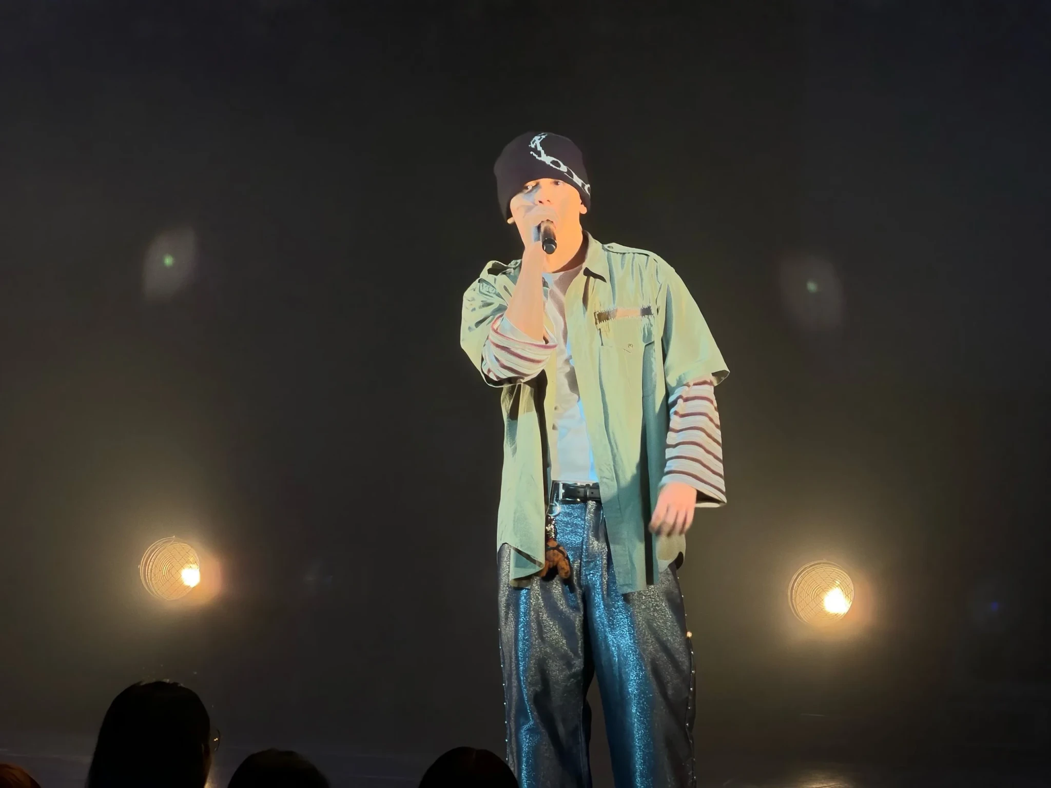 NEO of ONE OR EIGHT sings in a beanie and green overshirt with stage lights glowing behind him