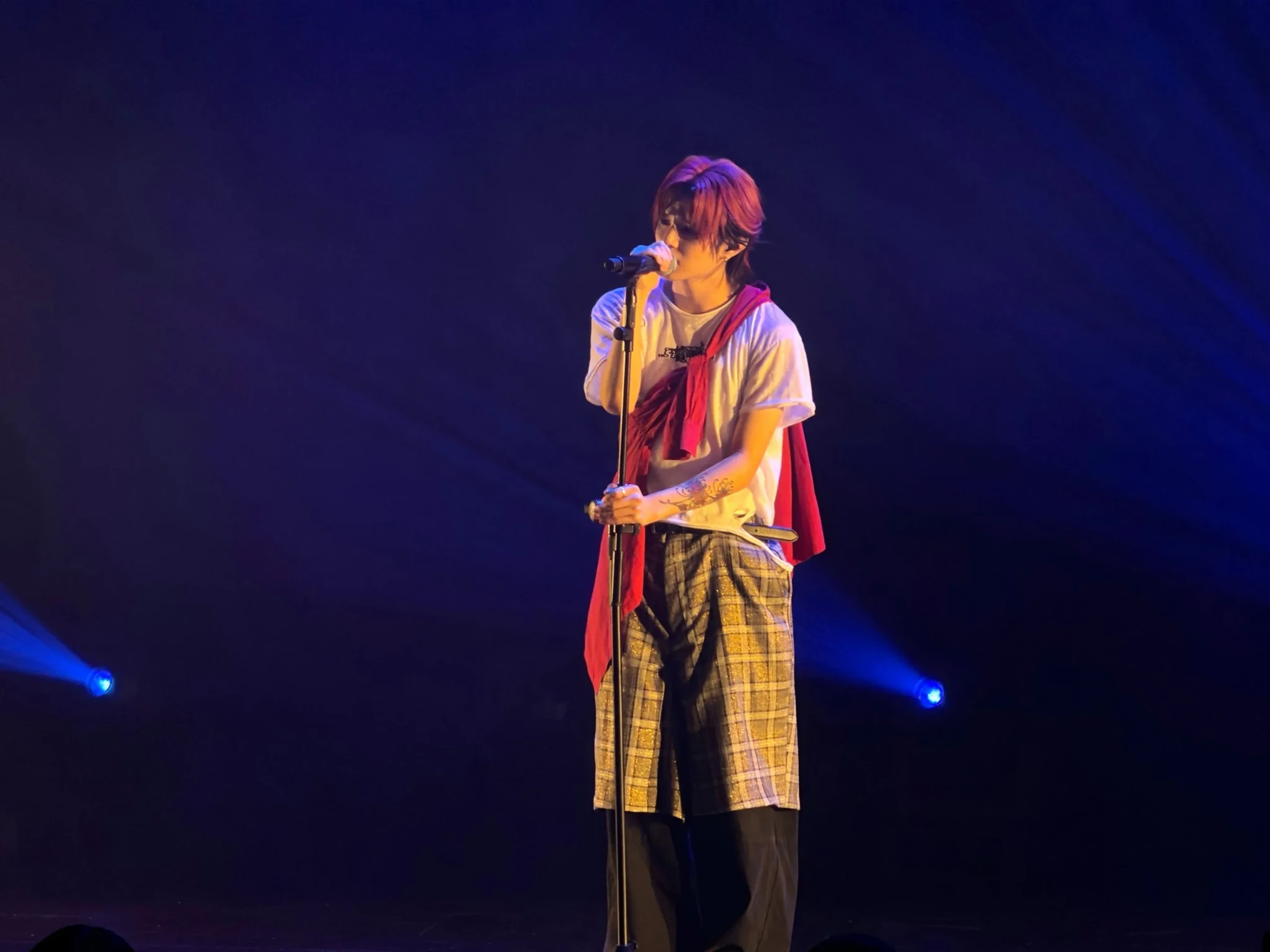 SOUMA of ONE OR EIGHT sings at a microphone stand in a red top over a white tee and a plaid skirt