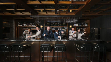 Five bartenders in suits and blazers working behind a dark marble bar, shaking cocktails and pouring spirits under a wooden coffered ceiling.