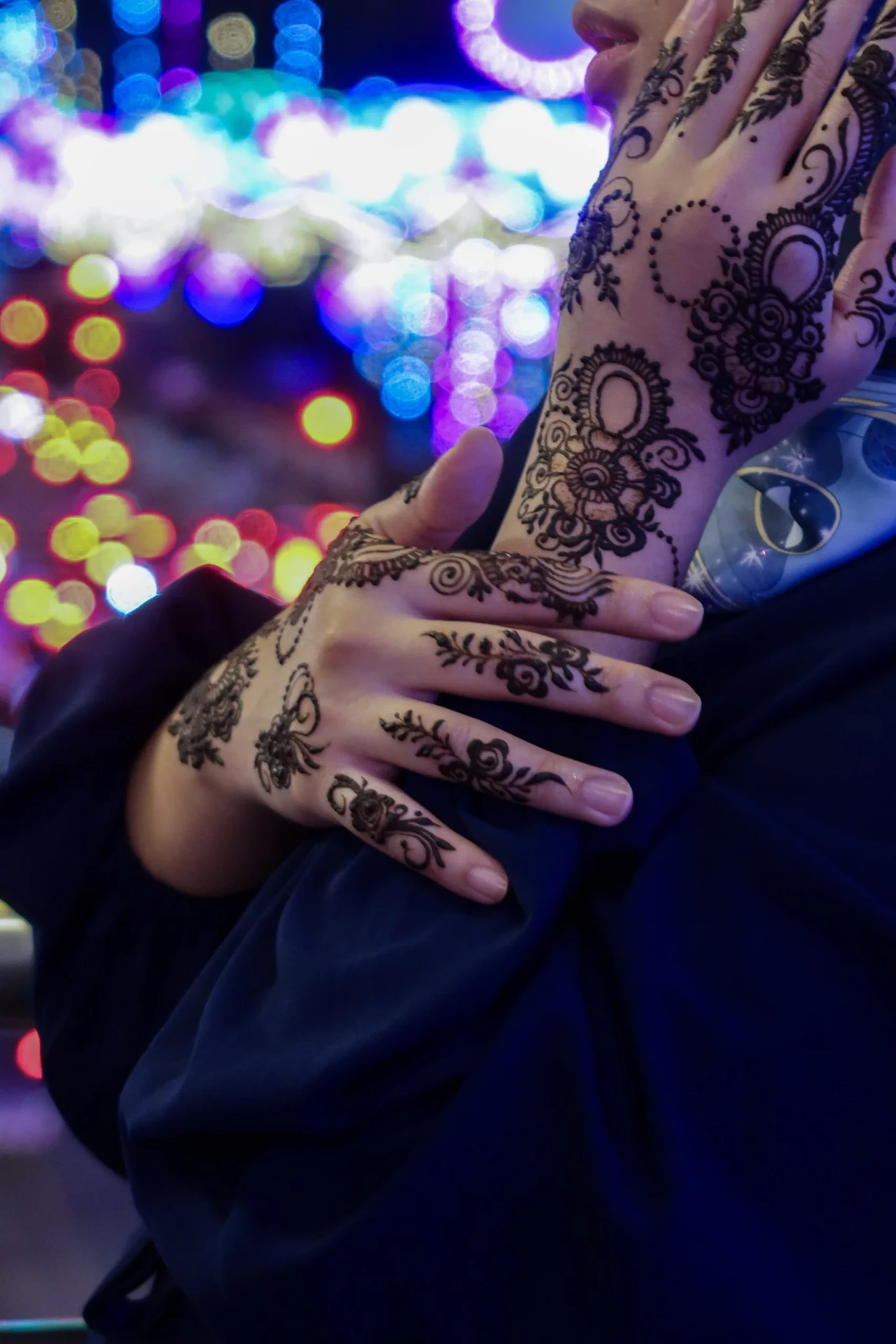 Hands decorated with black henna in floral and paisley motifs against blurred colourful lights