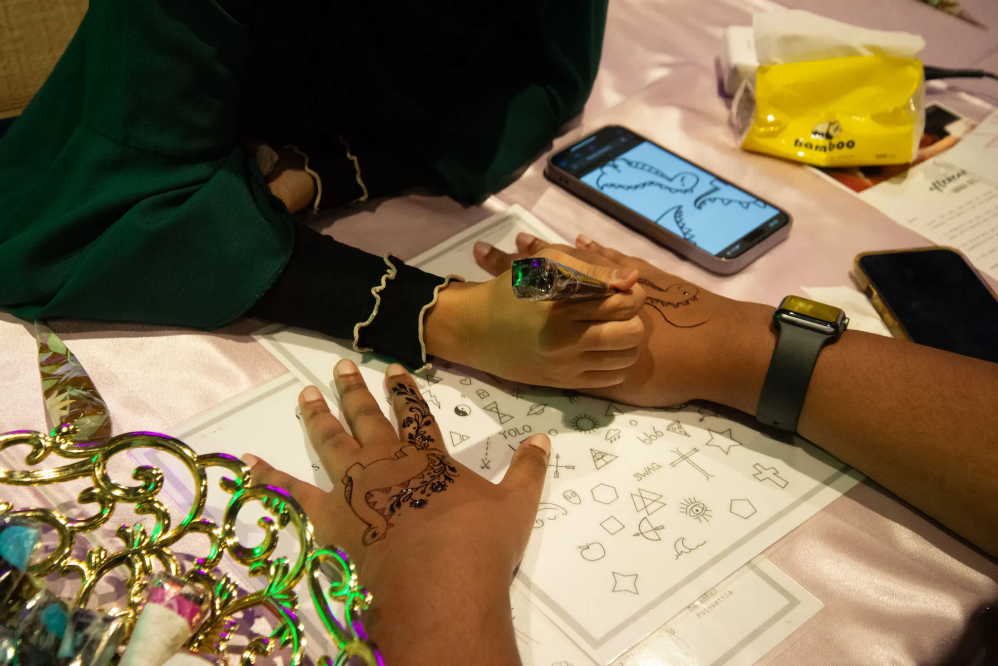Close-up of a henna design on a hand resting on a table beside a printed sheet of tattoo motifs and a phone showing a dinosaur drawing.