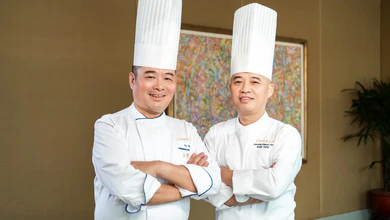 Chef Andy Fung and Chef Ku Keung in white chef's jackets and tall toques, posing with arms crossed