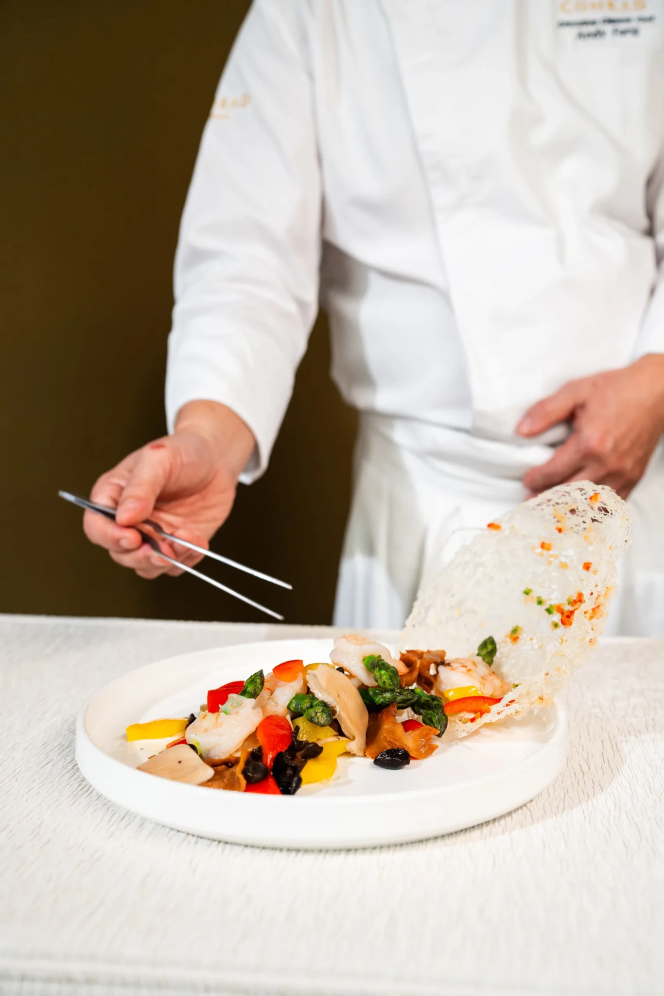Chef Andy Fung in a white chef's jacket using tweezers to garnish a plate of prawns, sea conch and peppers