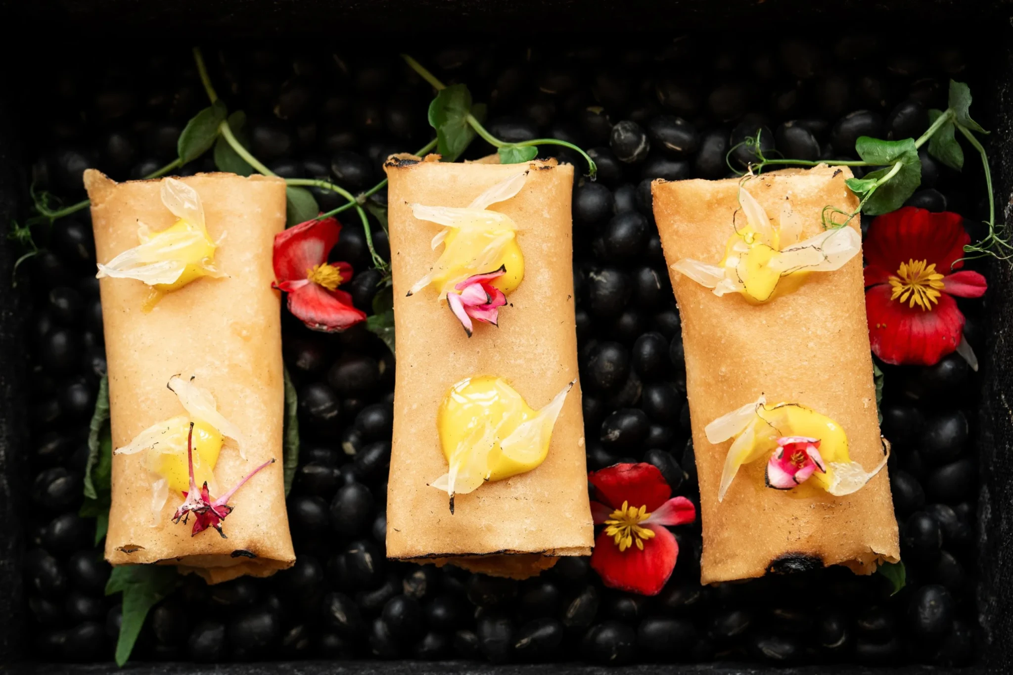 Golden sea cucumber spring rolls topped with yellow sauce and edible flowers on a bed of black beans