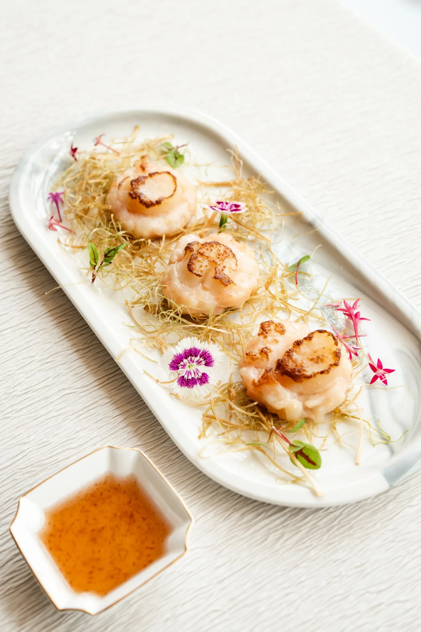 Pan-seared lemongrass cake with scallop on fried shredded threads and edible flowers, with amber sauce on a white plate