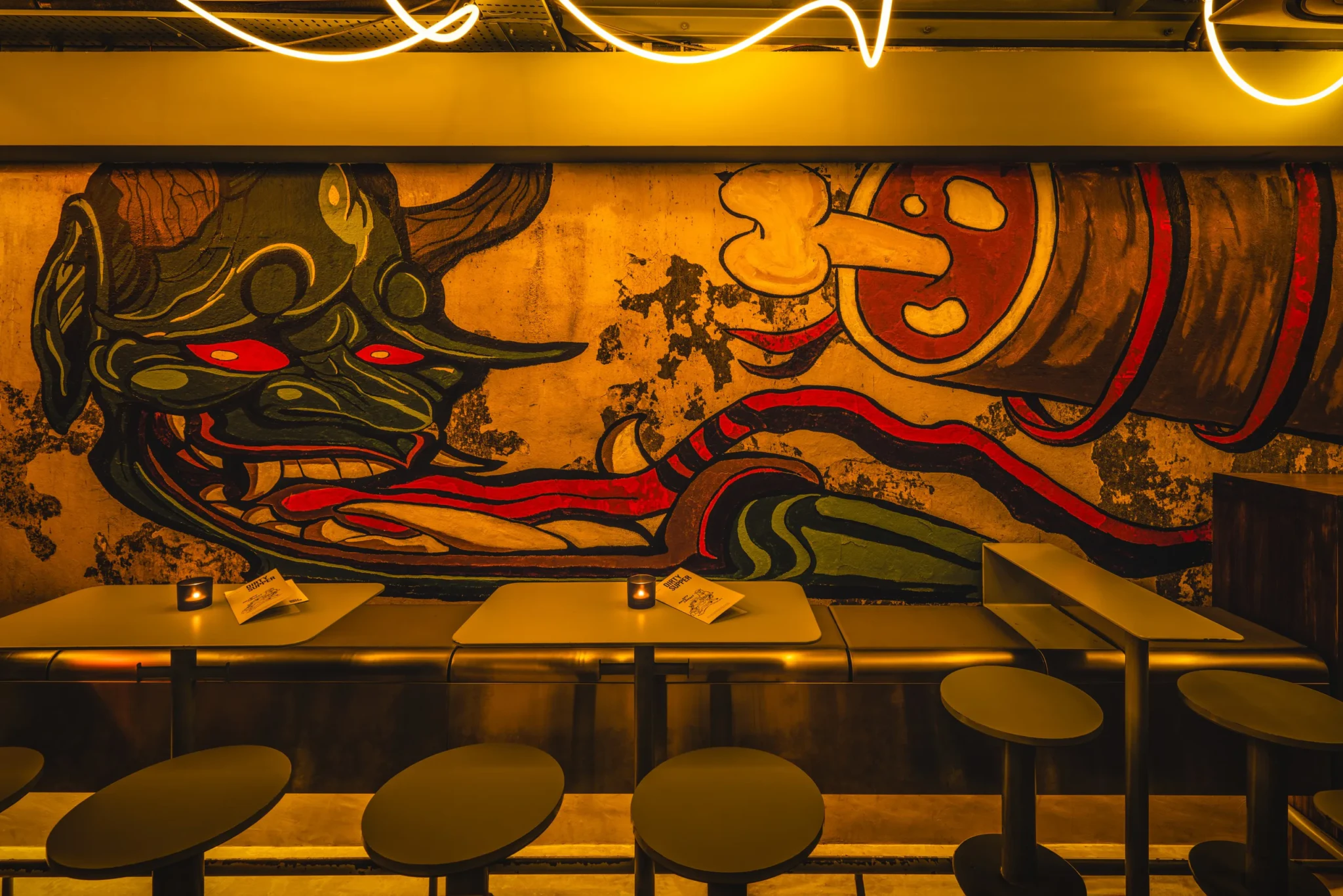 Bar interior with a painted demon mural on a weathered wall, round stools and small candles on tables.