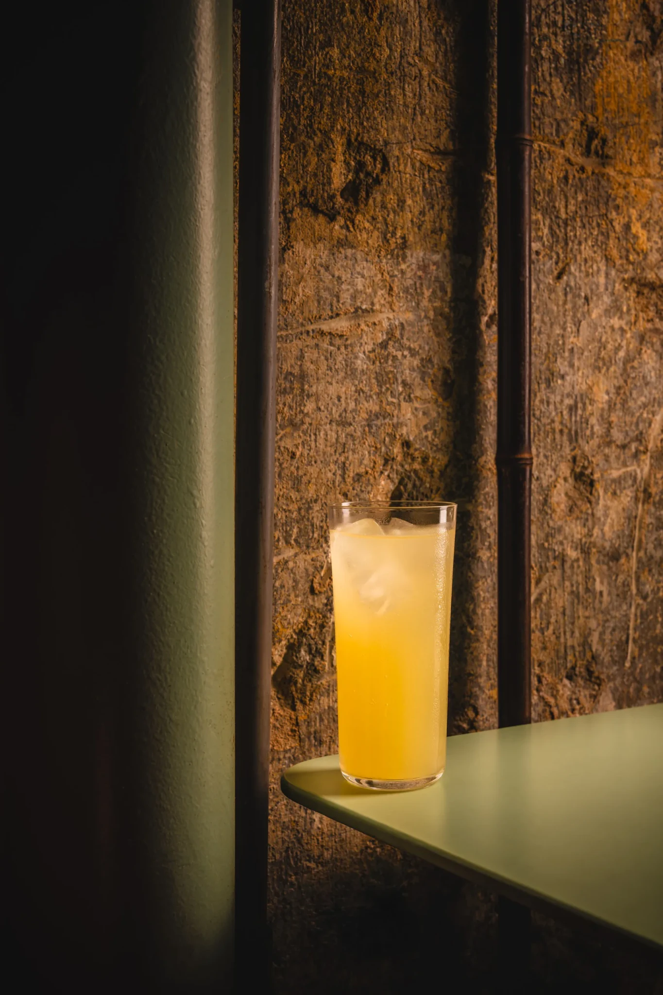 A tall glass of pale yellow cocktail over ice on a green shelf against a stone wall