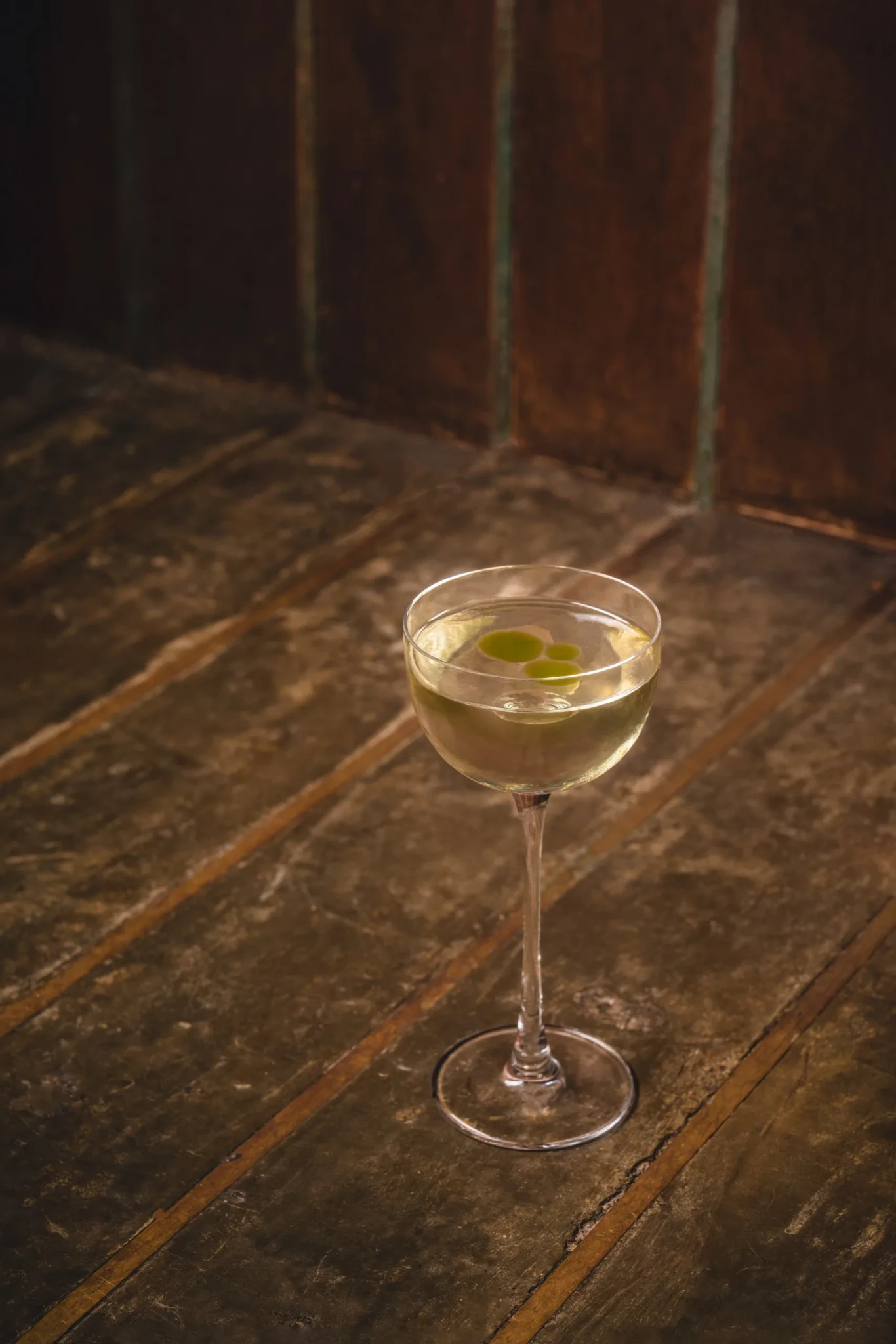 A chilled cocktail in a stemmed coupe glass with green oil droplets on the surface