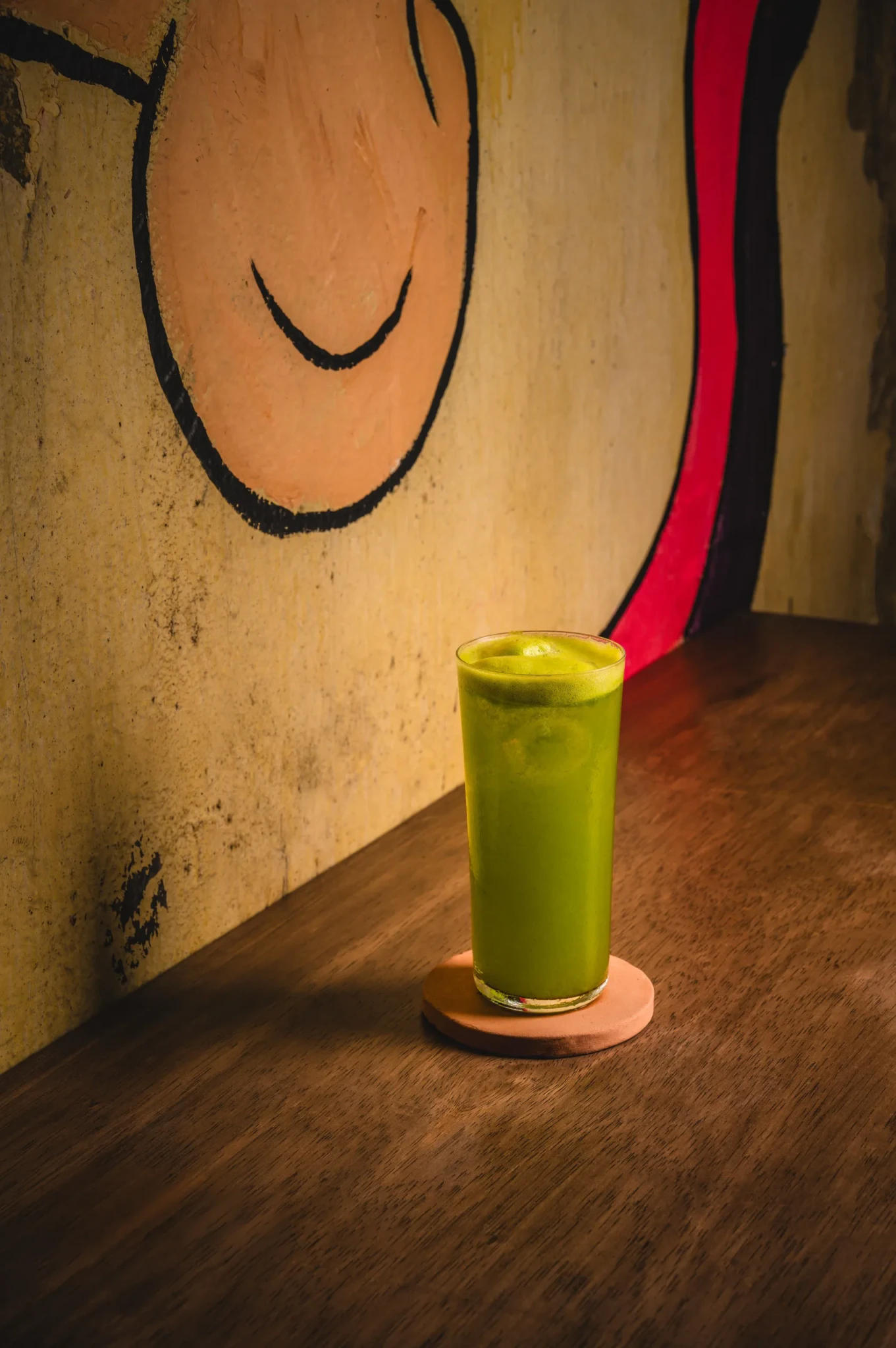 A bright green frothy cocktail in a tall glass on a wooden bar counter in front of a hand-painted mural