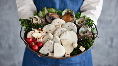 A server in a denim apron holds a metal tray of Purung Badang fishcake garnished with abalone, lettuce, cherry tomatoes and garlic