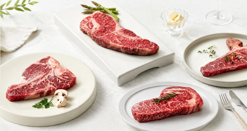 Cuts of marbled raw beef, including T-bone and ribeye, arranged on plates and a board with asparagus, mushrooms and herbs