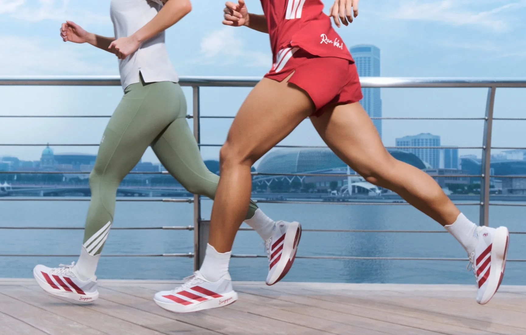 Two runners in the adidas SGIM26 capsule collection, including ADIZERO Evo SL shoes with red stripes and 'Singapore' printed on the sole, run along a waterfront