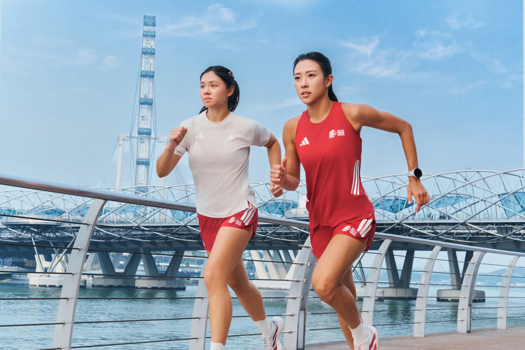 Two runners in adidas running apparel jog along a waterfront promenade with a Ferris wheel and arched footbridge behind them