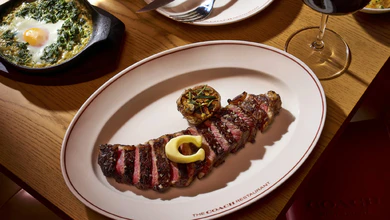 A sliced steak topped with a C-shaped pat of butter and roasted garlic on a plate printed with The Coach Restaurant logo