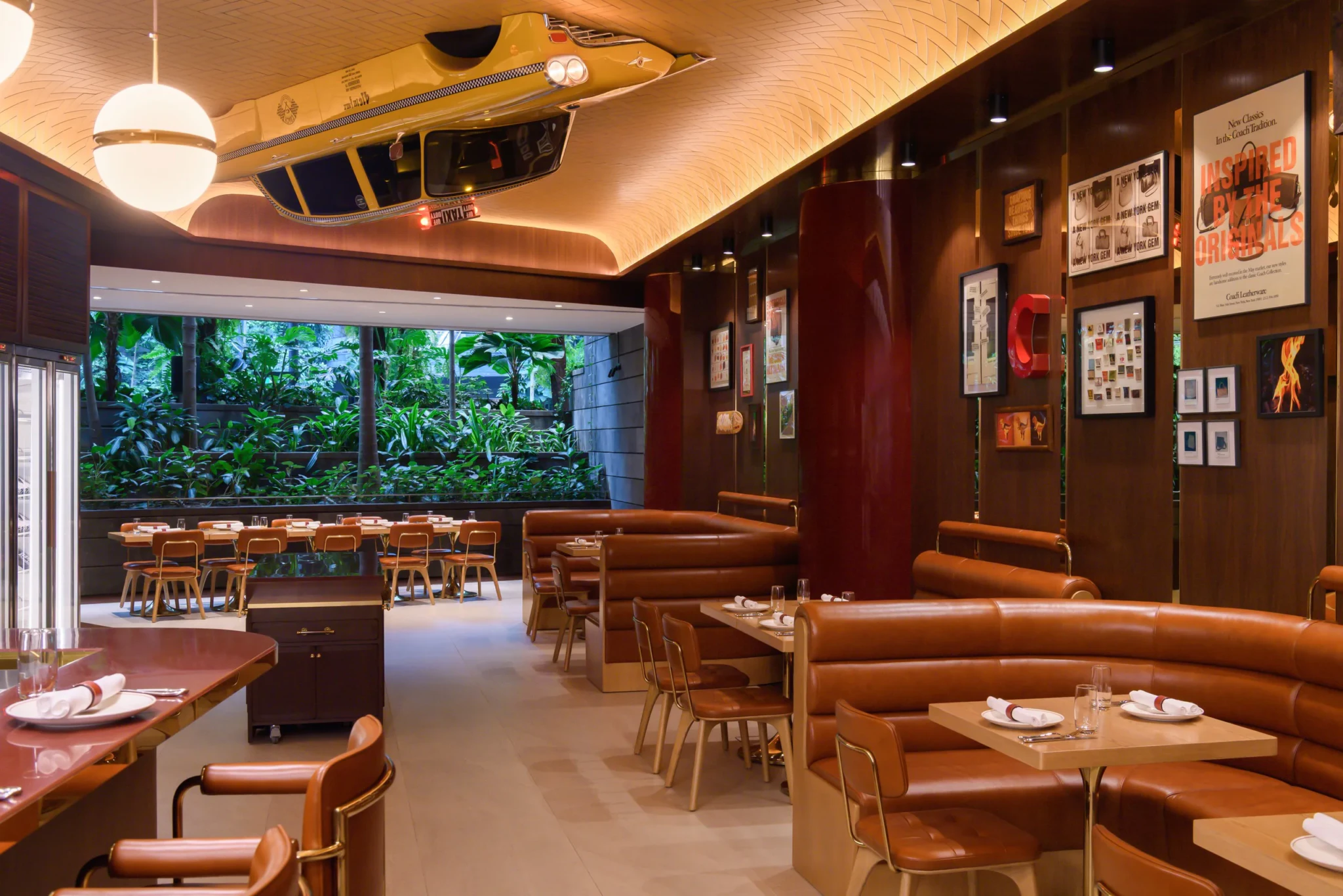 Interior of The Coach Restaurant with leather booths, a suspended yellow taxi, framed Coach ads and a view of greenery through floor-to-ceiling windows