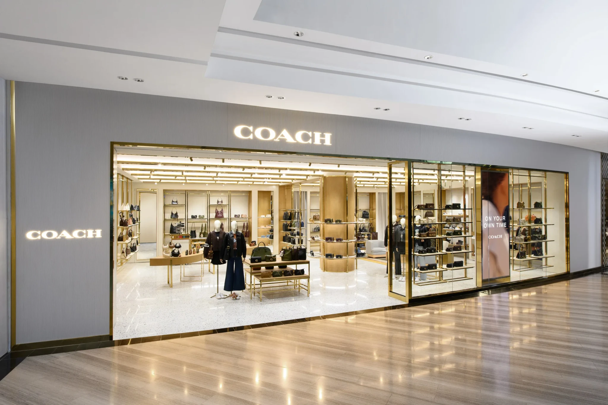 Coach boutique storefront with gold shelving, handbags on display, and mannequins in illuminated interior