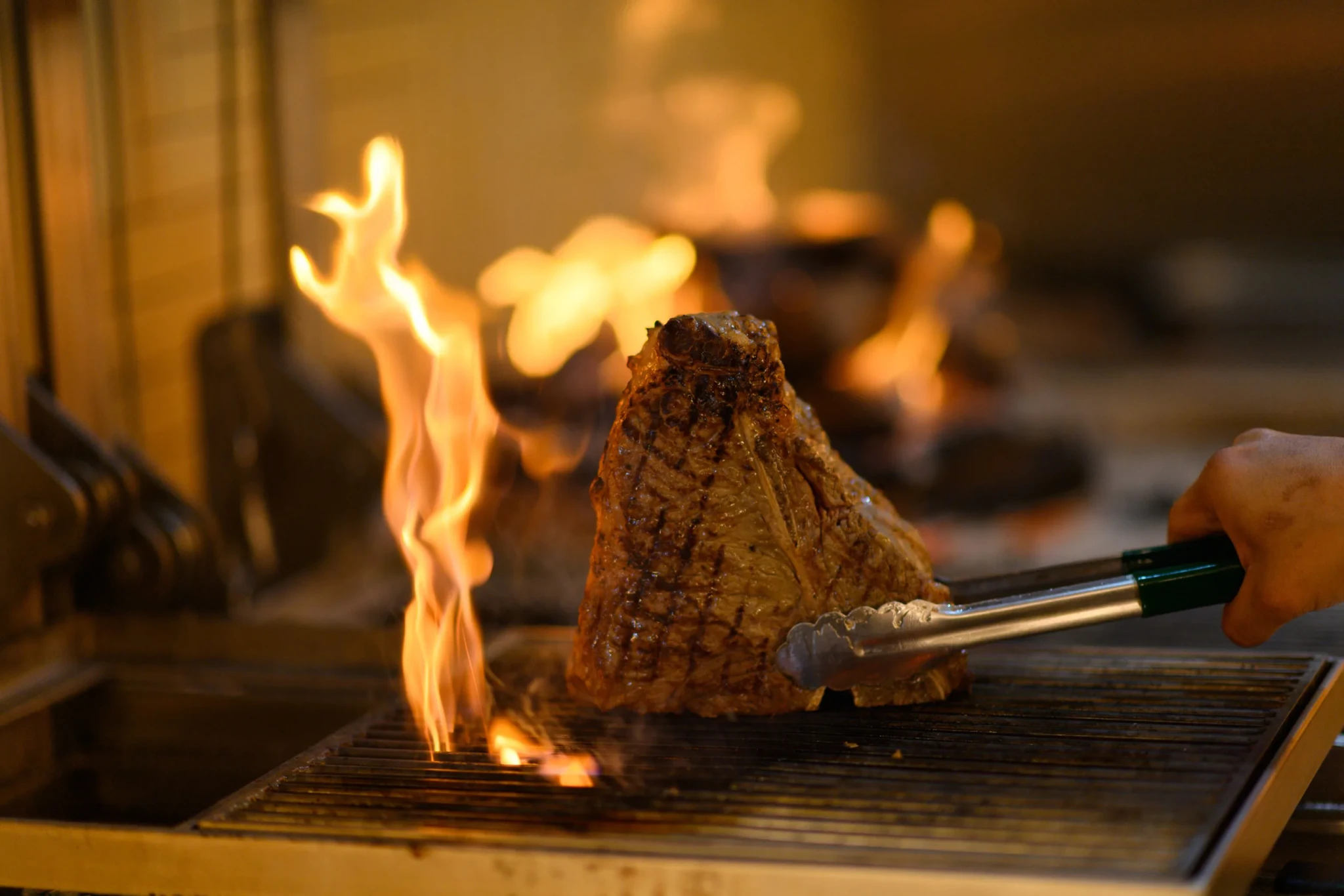 A chef turns a steak with tongs over an open flame at The Coach Restaurant