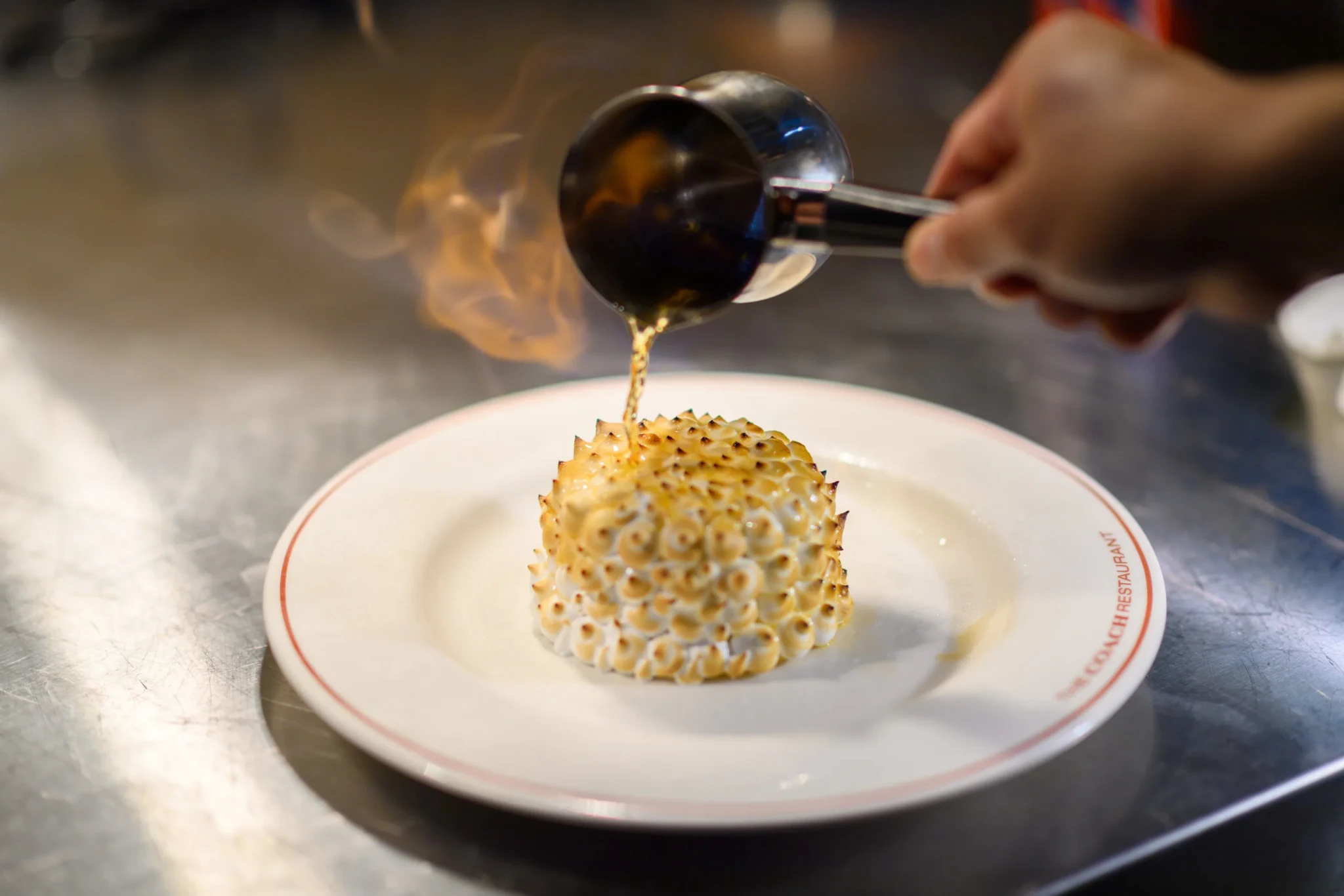 A chef pours flaming liquid over a meringue-covered Bombe Alaska on a plate printed The Coach Restaurant