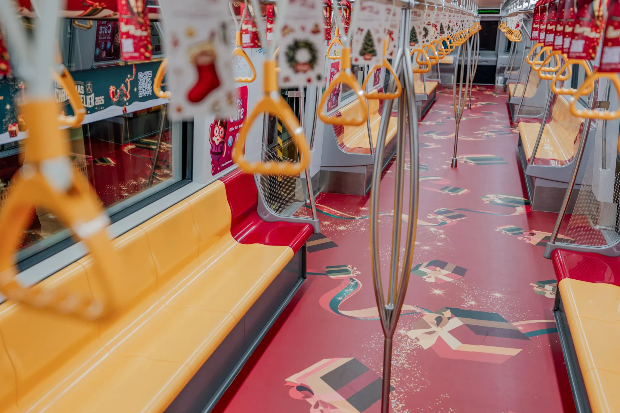 Interior of a train carriage decorated for The Christmas Atelier 2026, with red and yellow seats, festive gift-box floor graphics and hanging Christmas ornament
