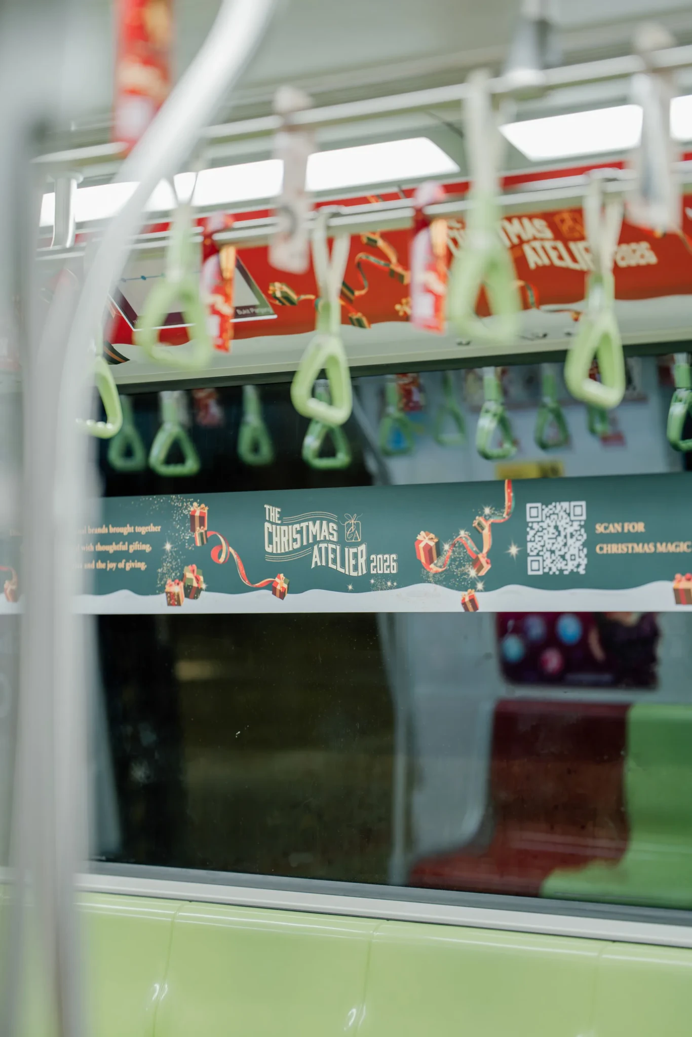 Interior of a train carriage decorated for The Christmas Atelier 2026, with green hand straps and a teal banner promoting the festive marketplace at Marina Bay 