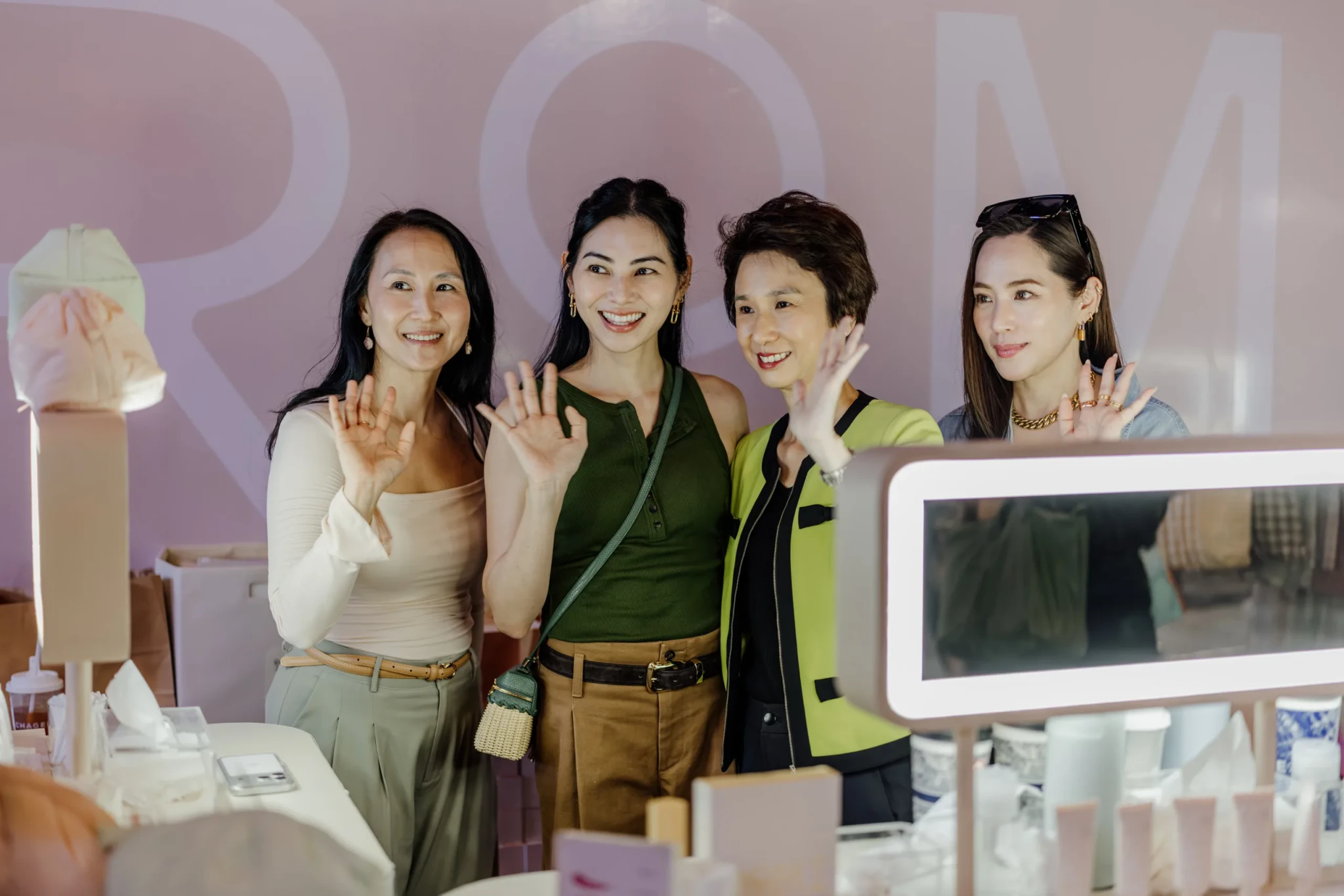 Four women smile and wave at a festive marketplace booth in front of a pink wall, with a lighted mirror and cosmetics in the foreground