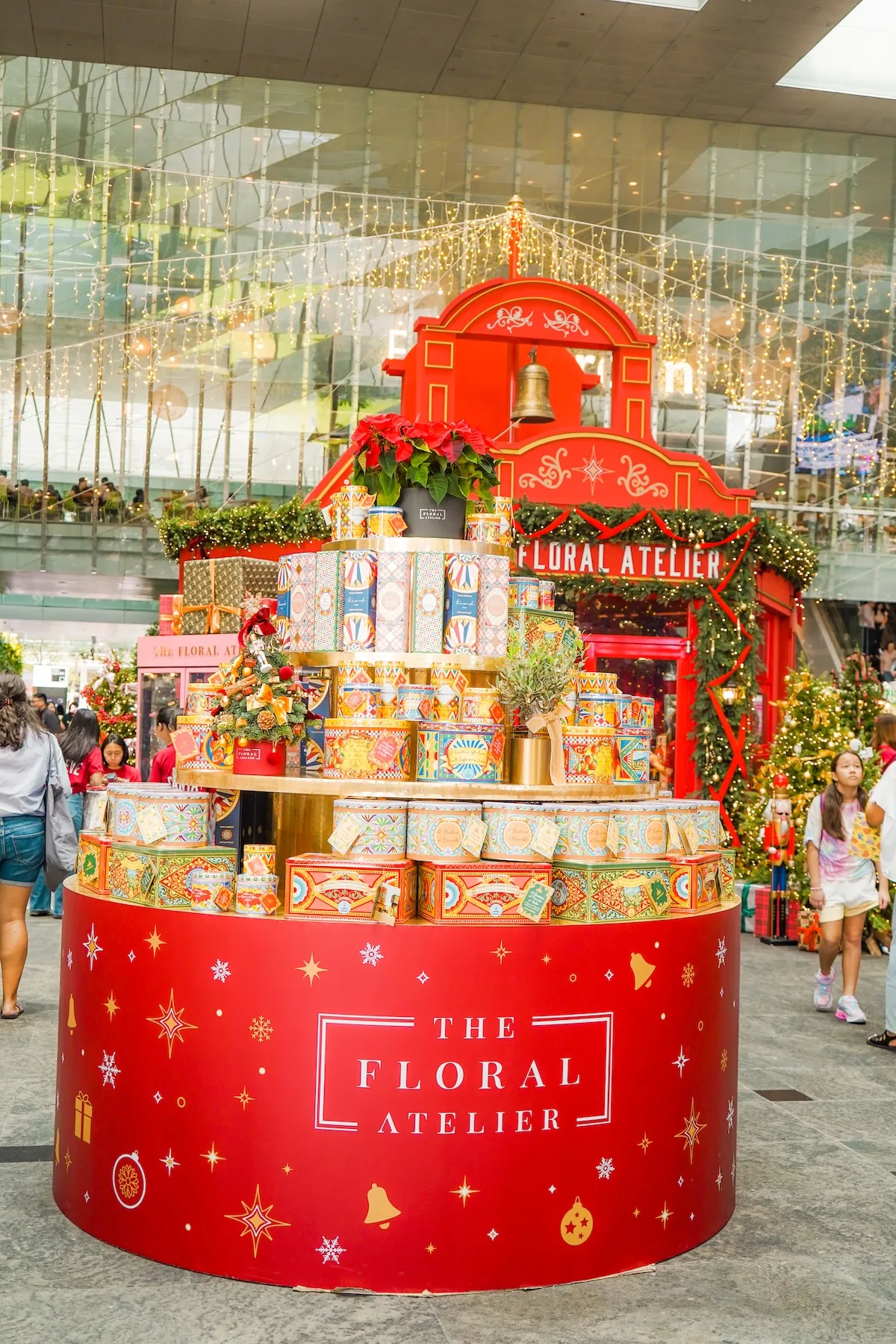 The Floral Atelier's festive display with stacked patterned gift tins and a poinsettia in front of a red holiday kiosk under string lights