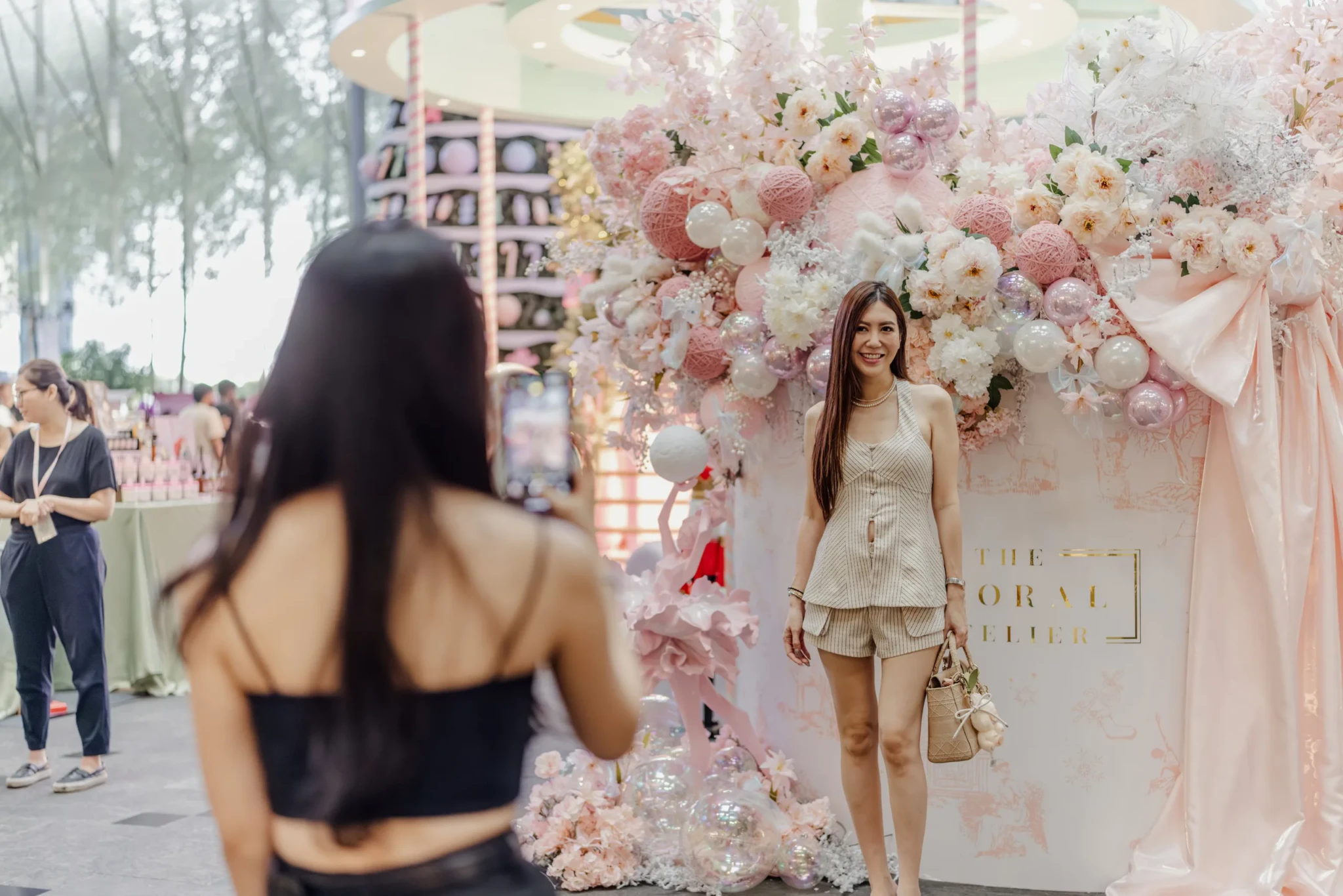 A woman in a striped beige set smiles in front of a pastel-pink floral installation with a sheer bow while another guest photographs her on a phone