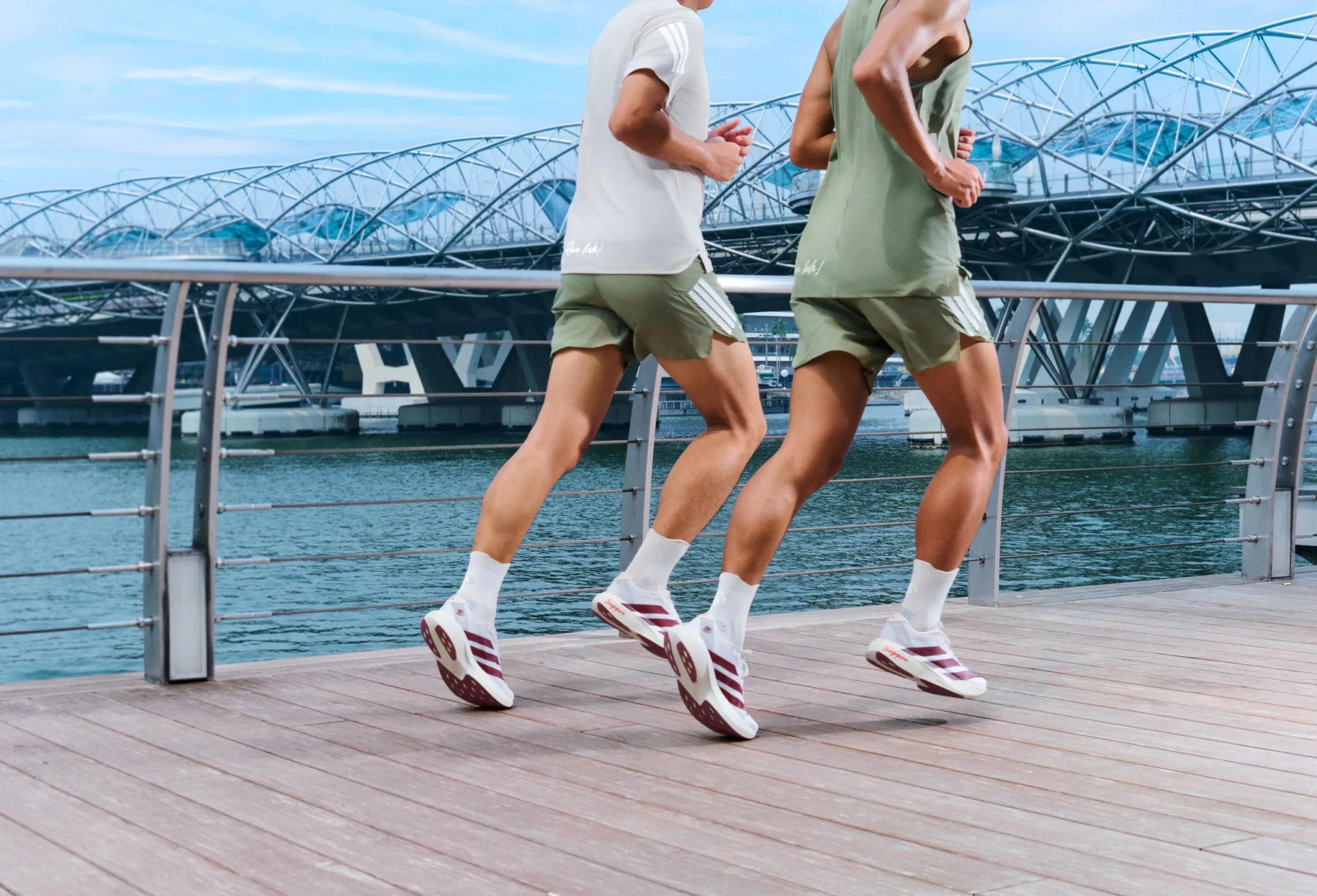 Two runners in adidas apparel and white-and-maroon running shoes jogging along a waterfront boardwalk beside a steel arch bridge