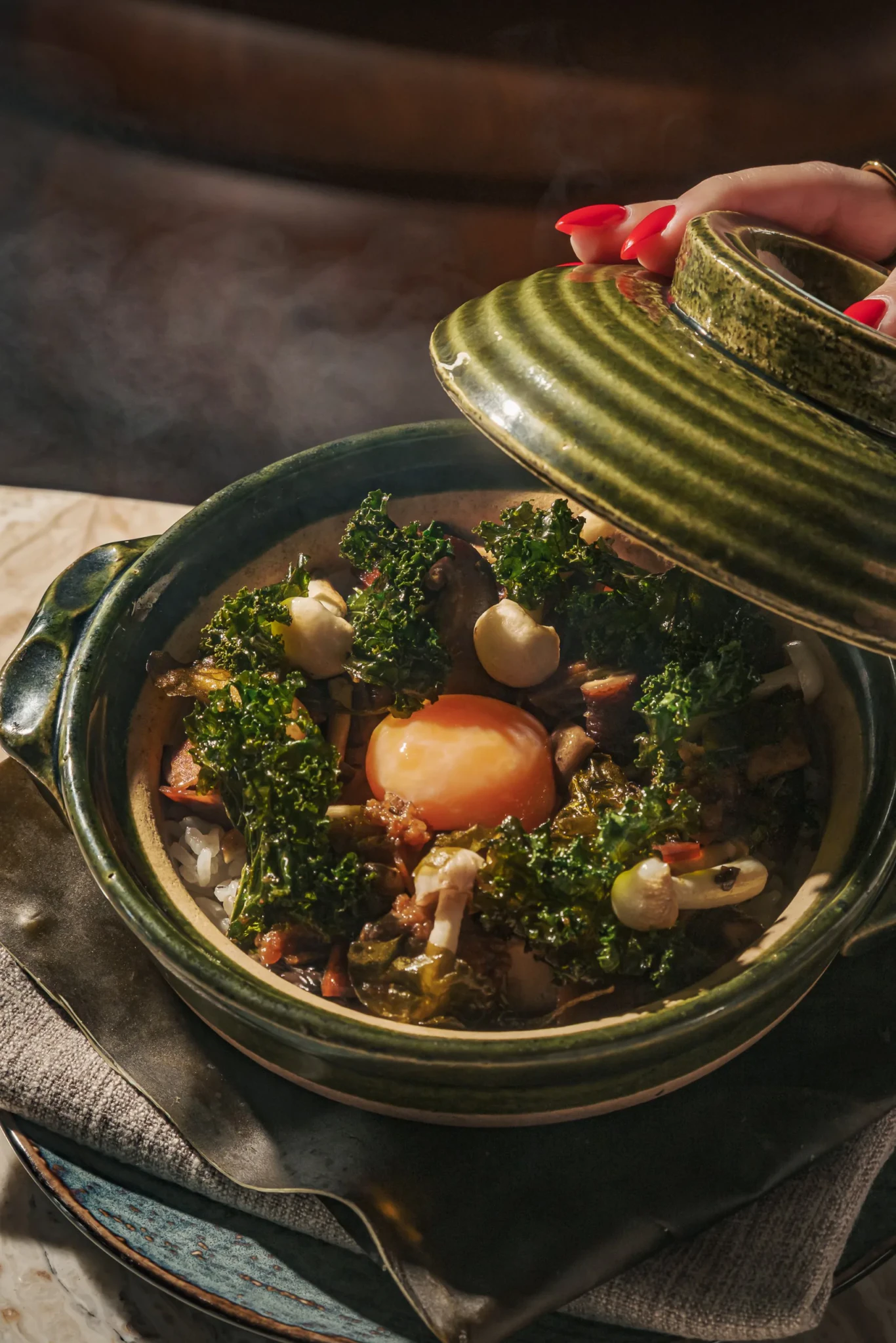 A hand lifts a green ceramic lid off a claypot of rice topped with kale, mushrooms and a raw egg yolk at CÉ LA VI Singapore